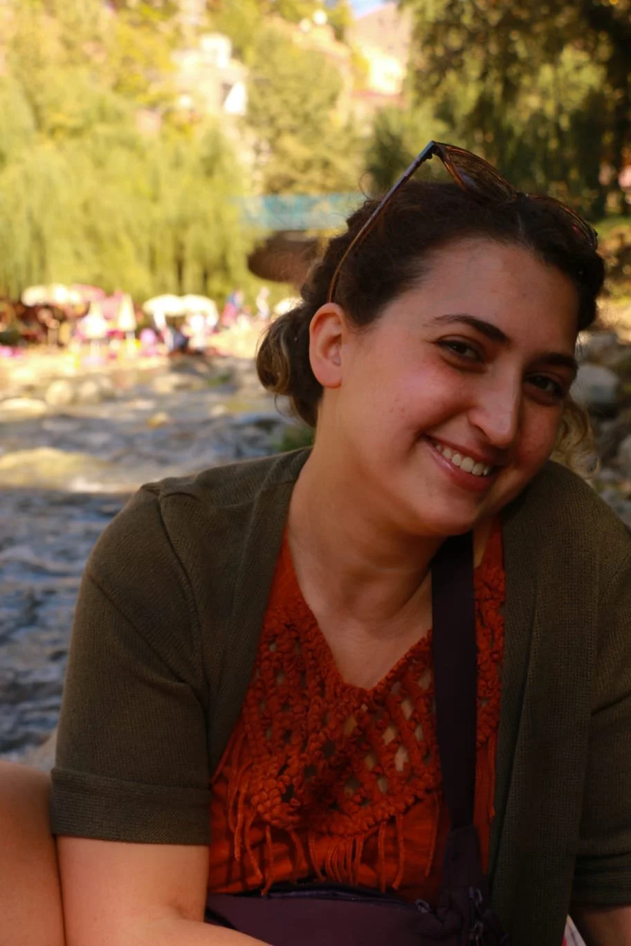 A smiling woman with sunglasses on her head sitting outdoors near a river, with trees and what appears to be a park in the background on a sunny day.
