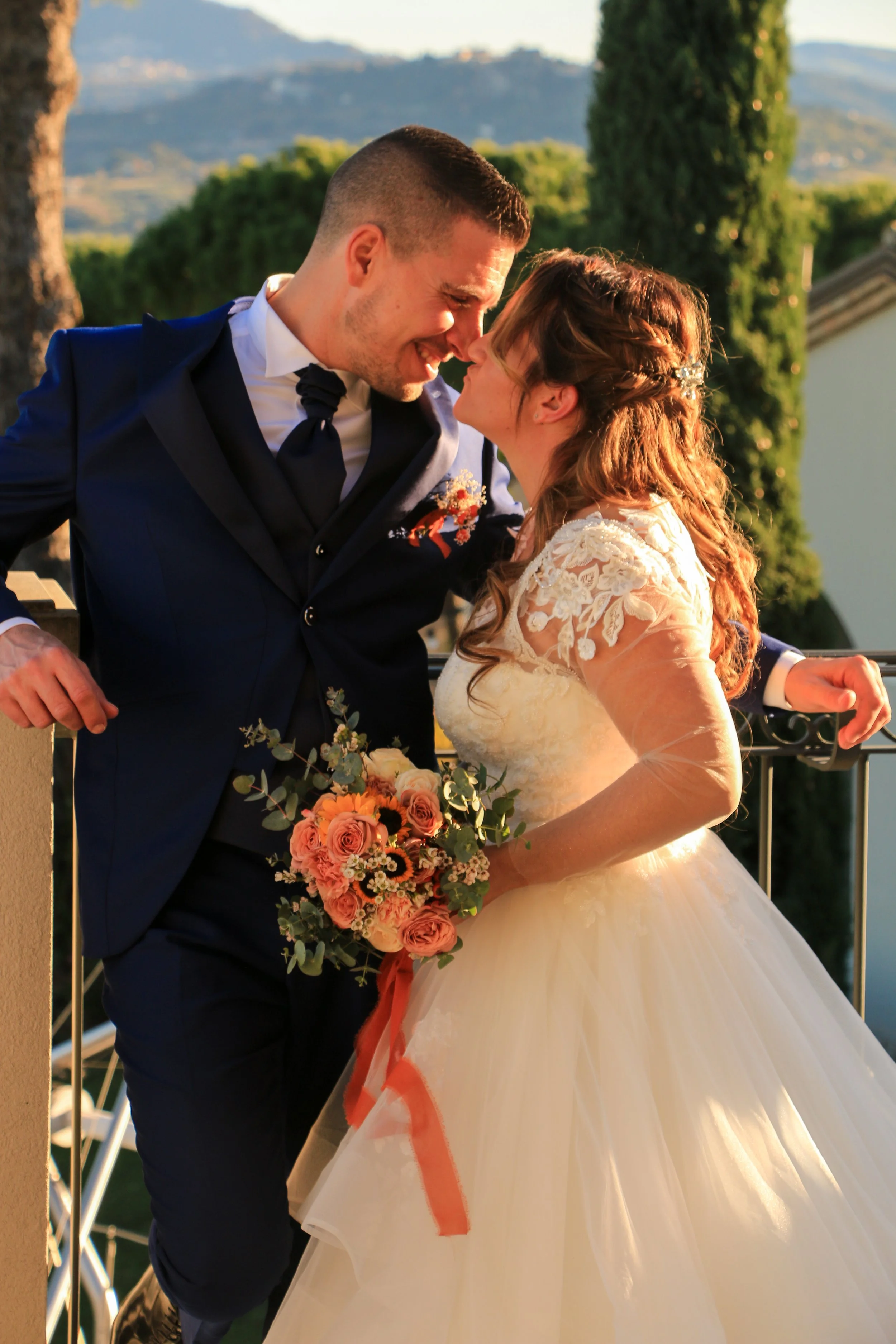 A bride and groom sharing a kiss outdoors during sunset, with greenery and mountains in the background. The bride is wearing a white wedding gown and holding a bouquet of pink and peach roses, while the groom is in a dark suit.