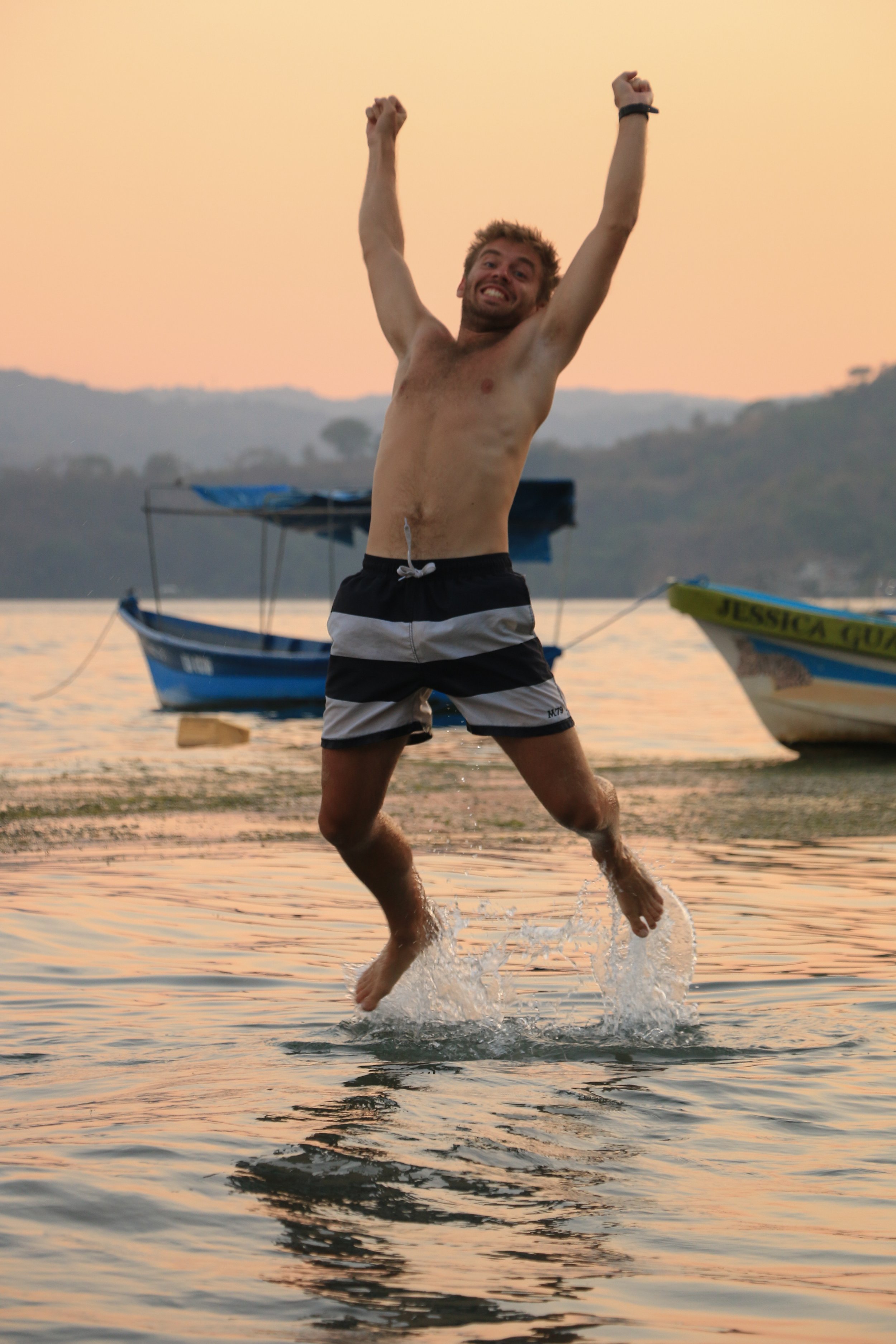 A man jumping into water during sunset with boats in the background.