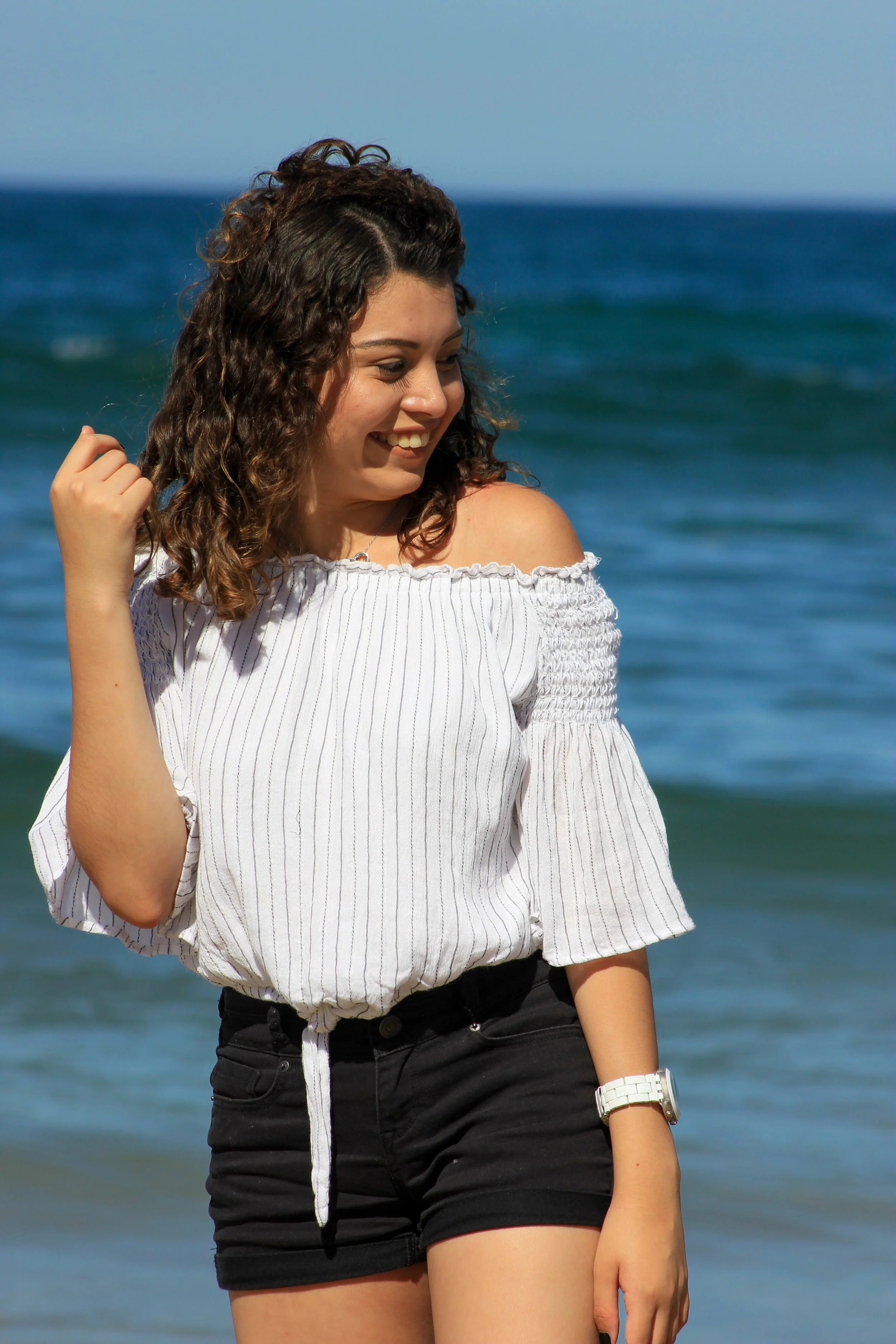 A young woman in a white off-shoulder top and black shorts standing by the ocean, smiling and looking down.