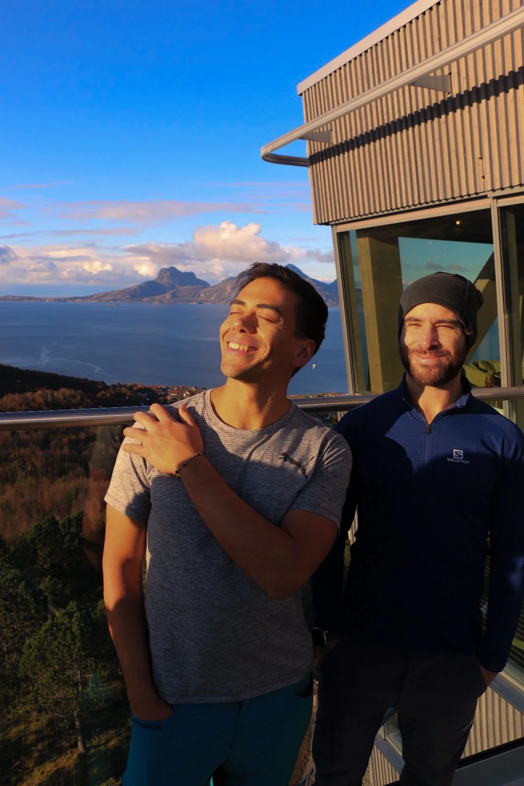 Two men smiling outdoors on a balcony with scenic mountain and water view in the background.