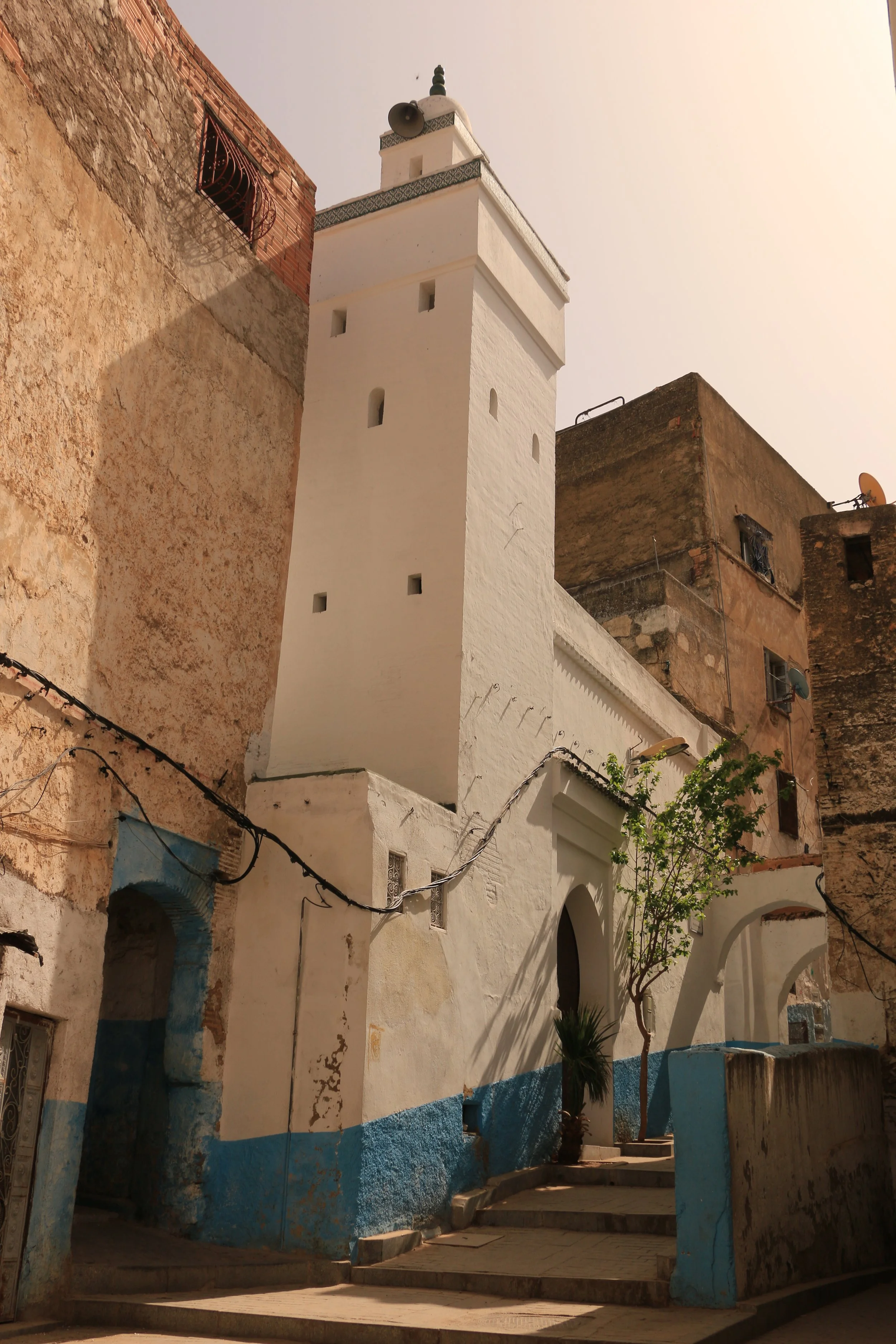 A white Moroccan-style minaret with small windows and a decorative top, flanked by weathered buildings with a blue and white base, a small tree, and steps leading to an arched doorway.