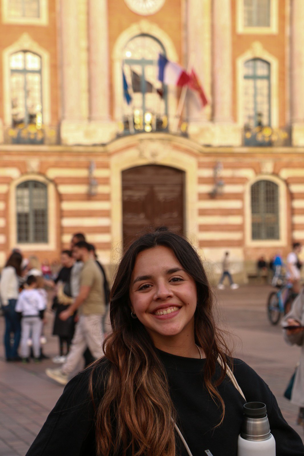 A young woman smiling in front of a historic brick building with flags and crowd.