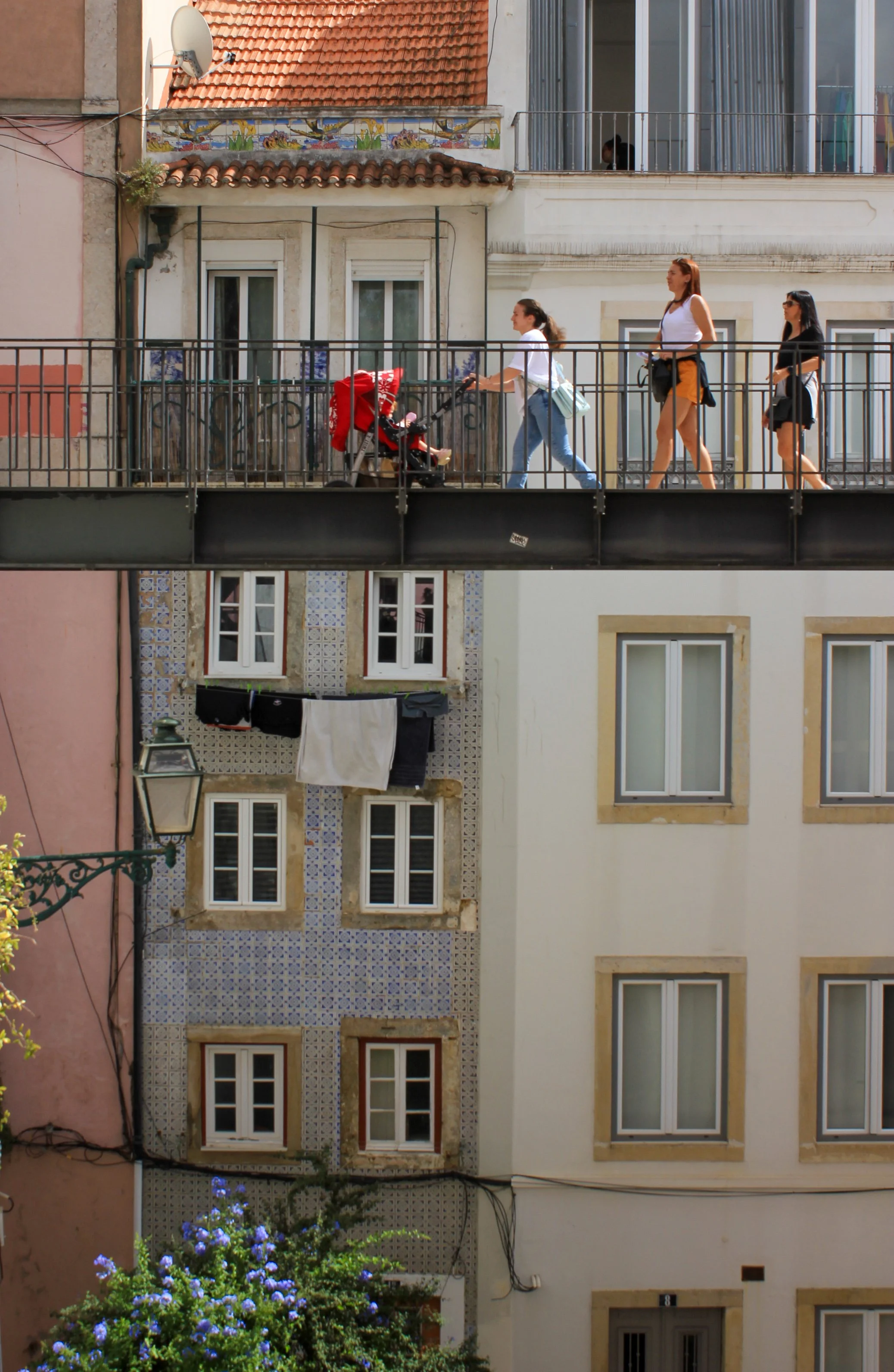 People walking on an elevated bridge over a city street, with multi-story buildings in the background and laundry hanging outside windows.