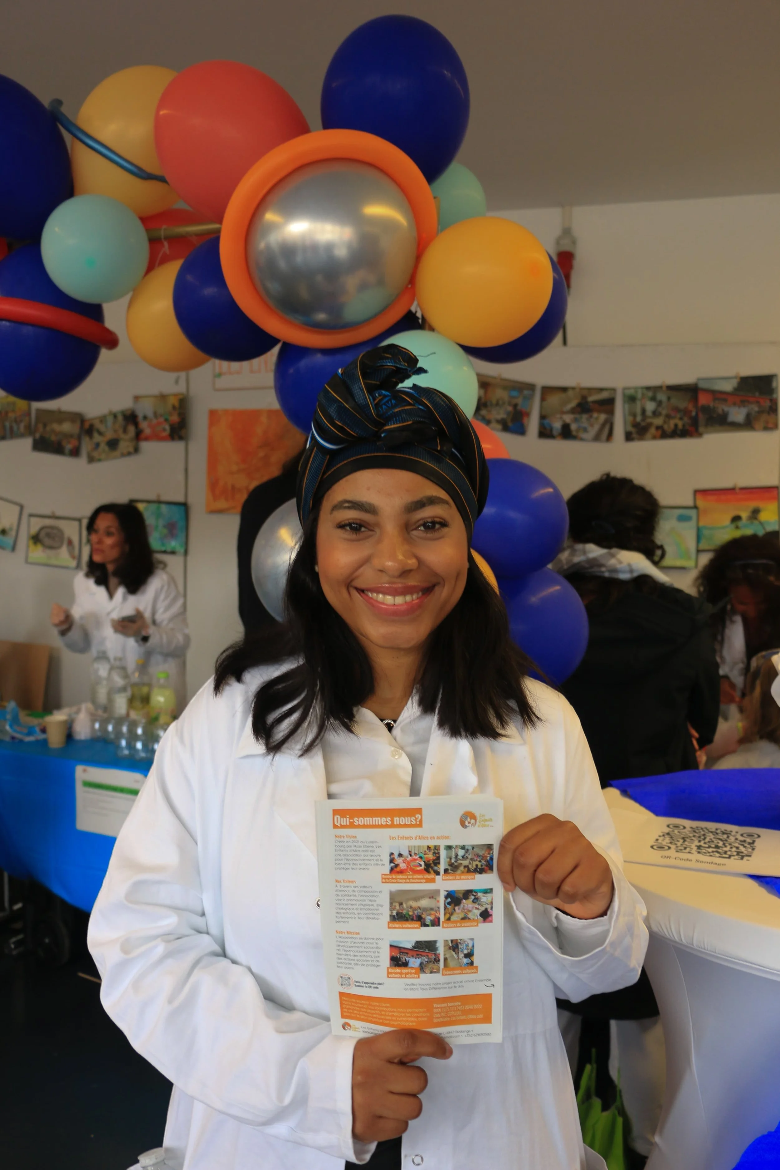 A woman with a headscarf and a white coat smiling and holding a flyer at an indoor event with balloons and other people in the background.