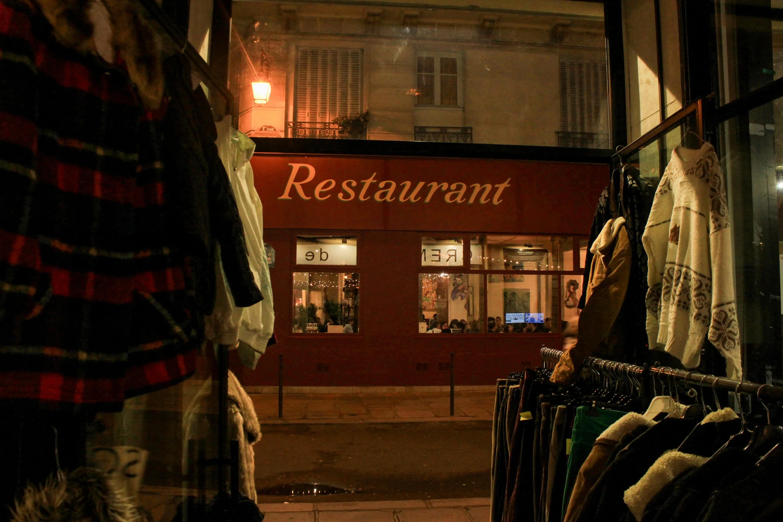 Night view of the exterior of a restaurant seen through a clothing shop window, with clothing racks and jackets in the foreground, and a streetlamp and building windows in the background.