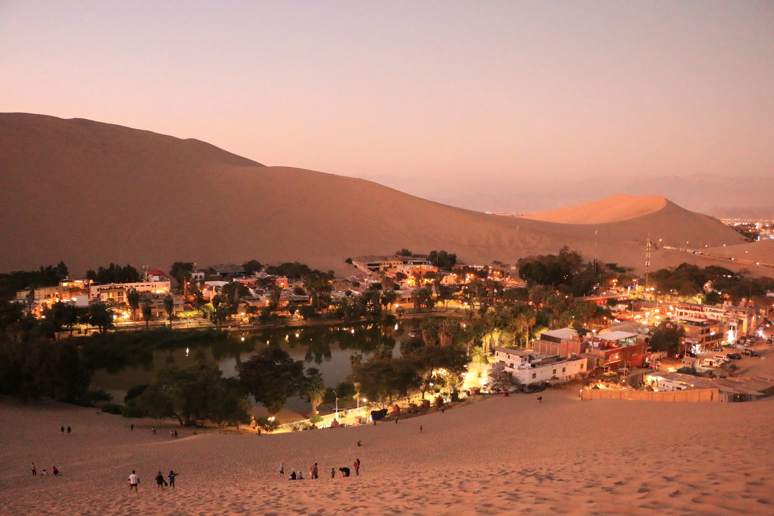 Dusk view of a town near the desert, with sand dunes in the background and a small illuminated pond surrounded by trees in the foreground.