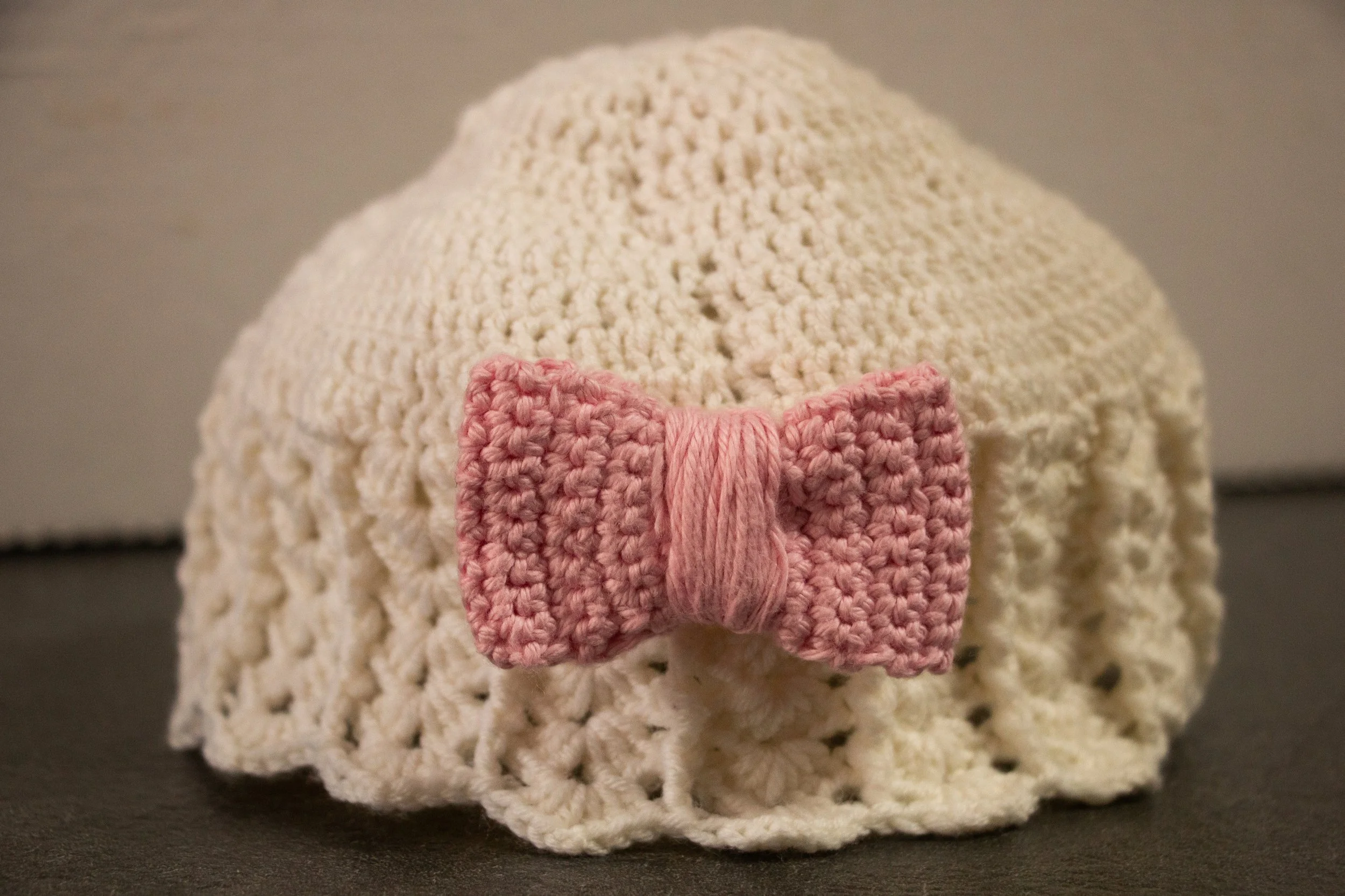 A crocheted cream-colored baby hat with a pink crocheted bow on the front.
