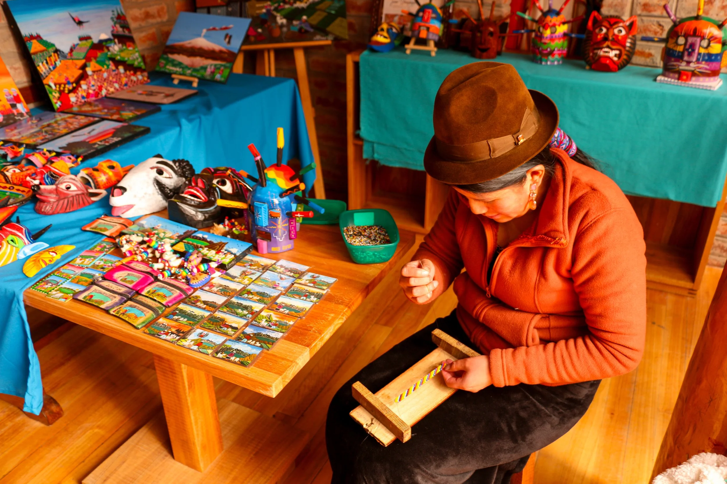 A woman wearing an orange jacket and a brown hat is sitting on a wooden bench, working on a colorful bead bracelet with a tool in her hand. She is in an artisan shop filled with colorful handcrafted masks, jewelry, paintings, and figurines displayed on tables with bright blue tablecloths.