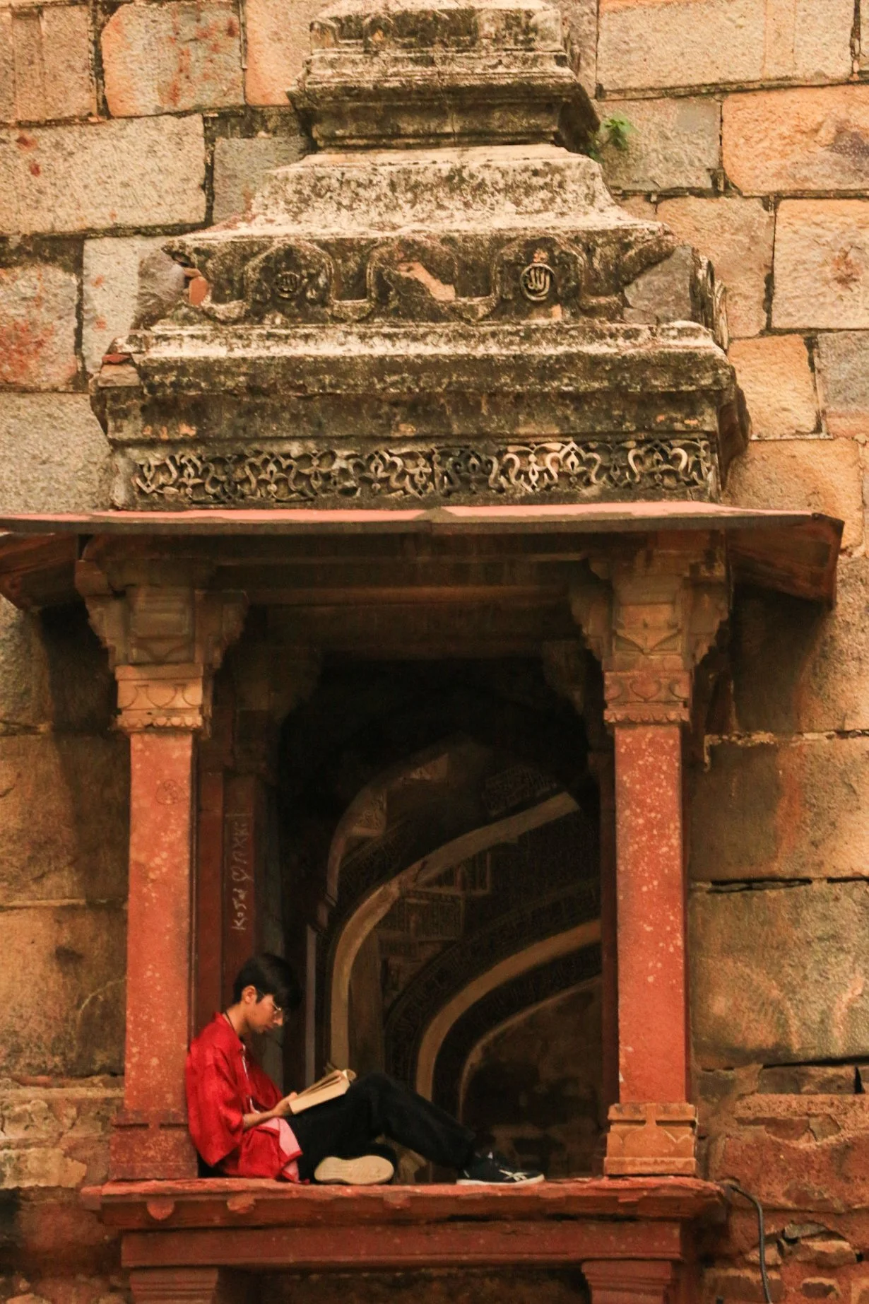 A young boy in a red shirt sitting on a stone ledge, reading a book, with an ancient stone and brick structure with carved details behind him.