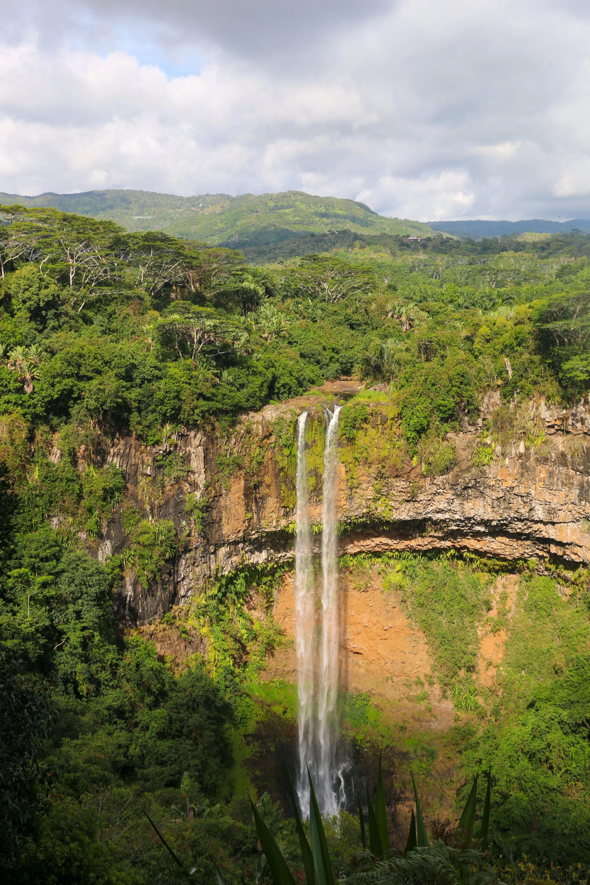 A tall waterfall flowing over a cliff into a lush green tropical jungle landscape with mountains in the background.