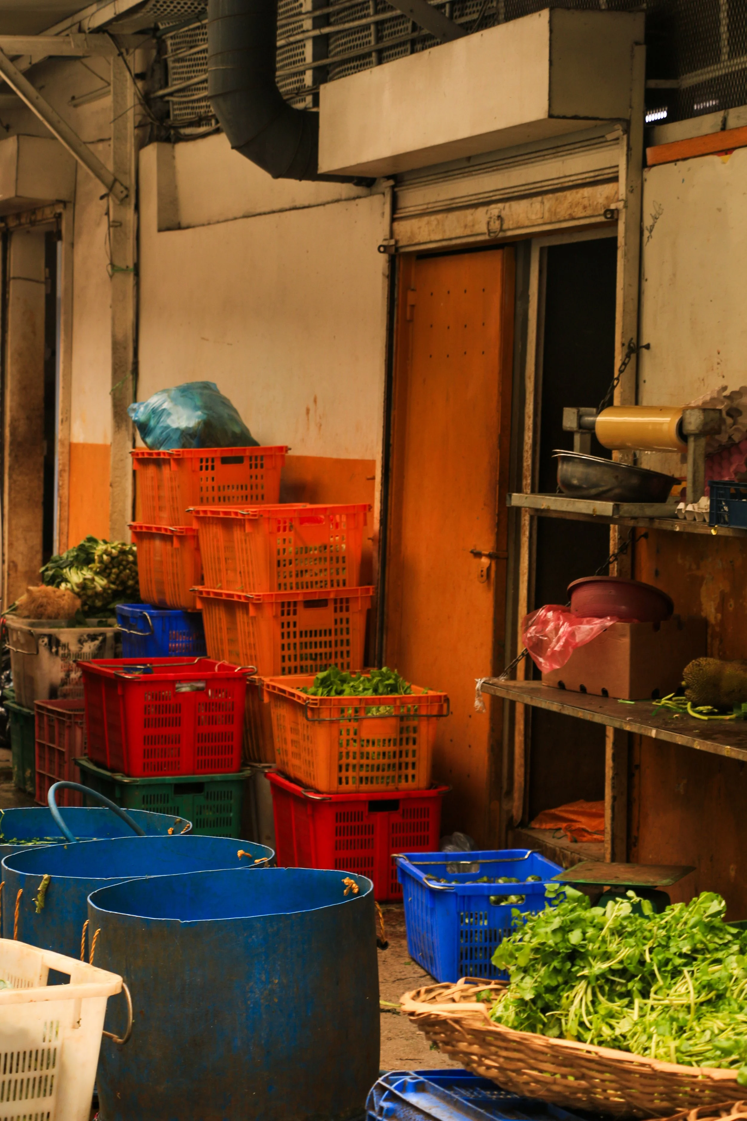 A street market scene with stacked orange and red plastic baskets, some filled with vegetables, and a blue basket in the foreground, along with a wicker basket of greens, against a backdrop of an aged storefront with a closed wooden door.