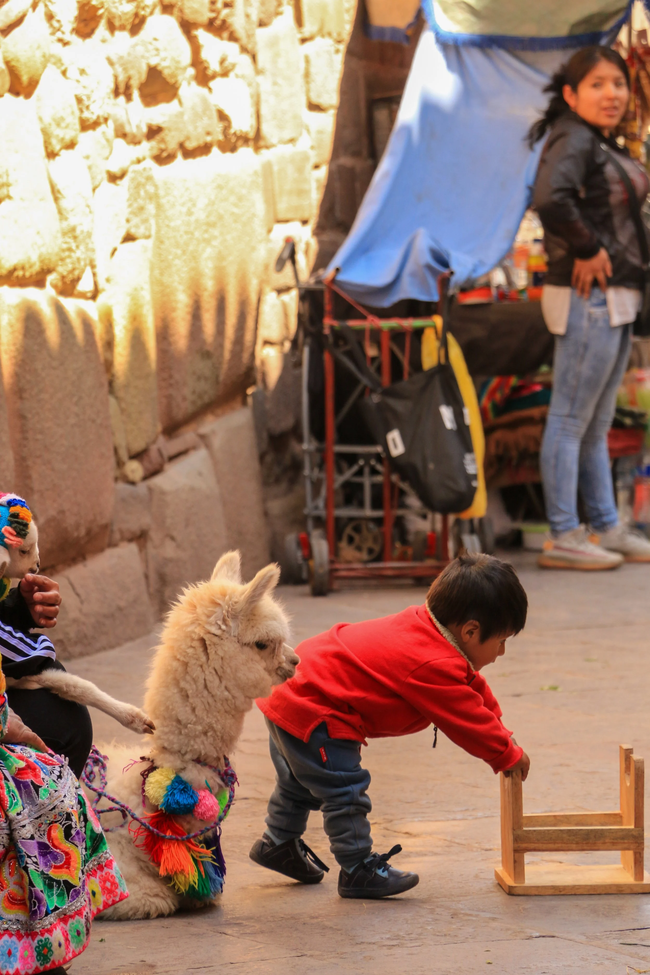 A young boy is playing with a small wooden chair on cobblestone street. A llama, decorated with colorful accessories, is standing nearby. A woman and a street vendor are in the background, along with a stone wall.