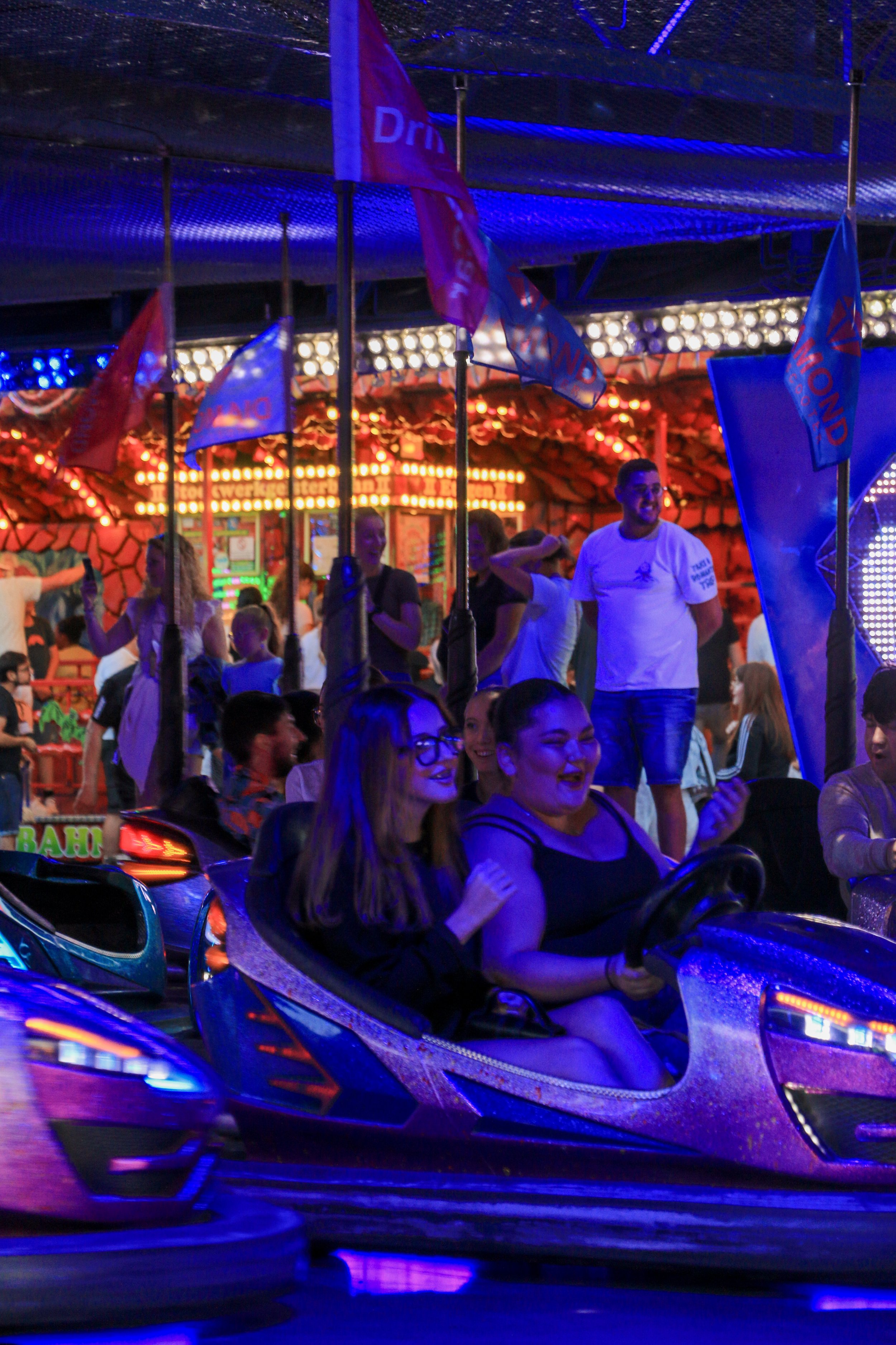 People riding bumper cars at a carnival or amusement park at night, with colorful lights and flags in the background.