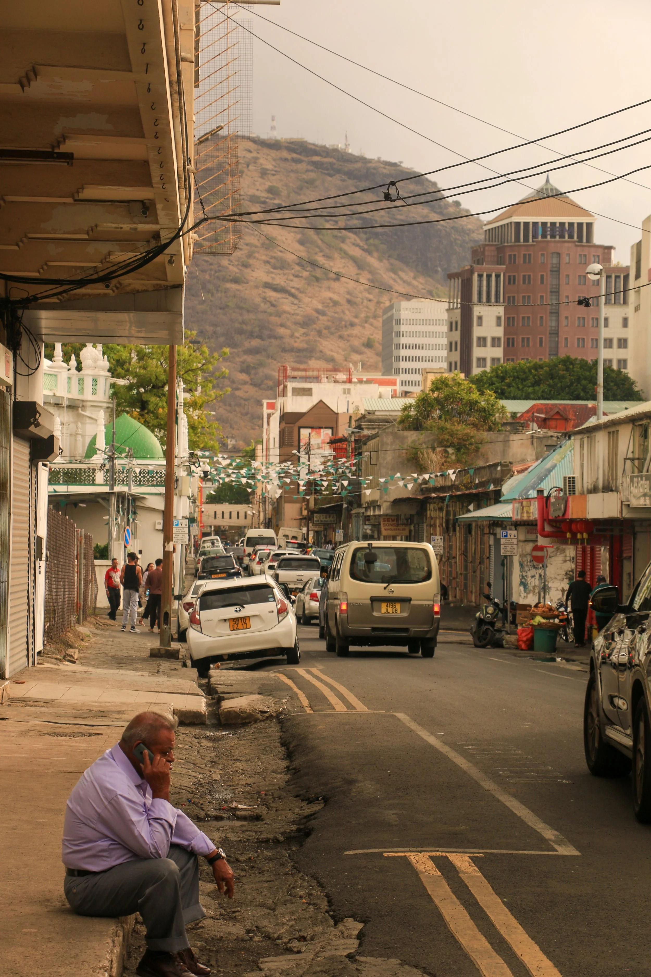 A man sitting on the sidewalk talking on his cell phone on a busy street with cars and pedestrians, with a mountain and modern buildings in the background.