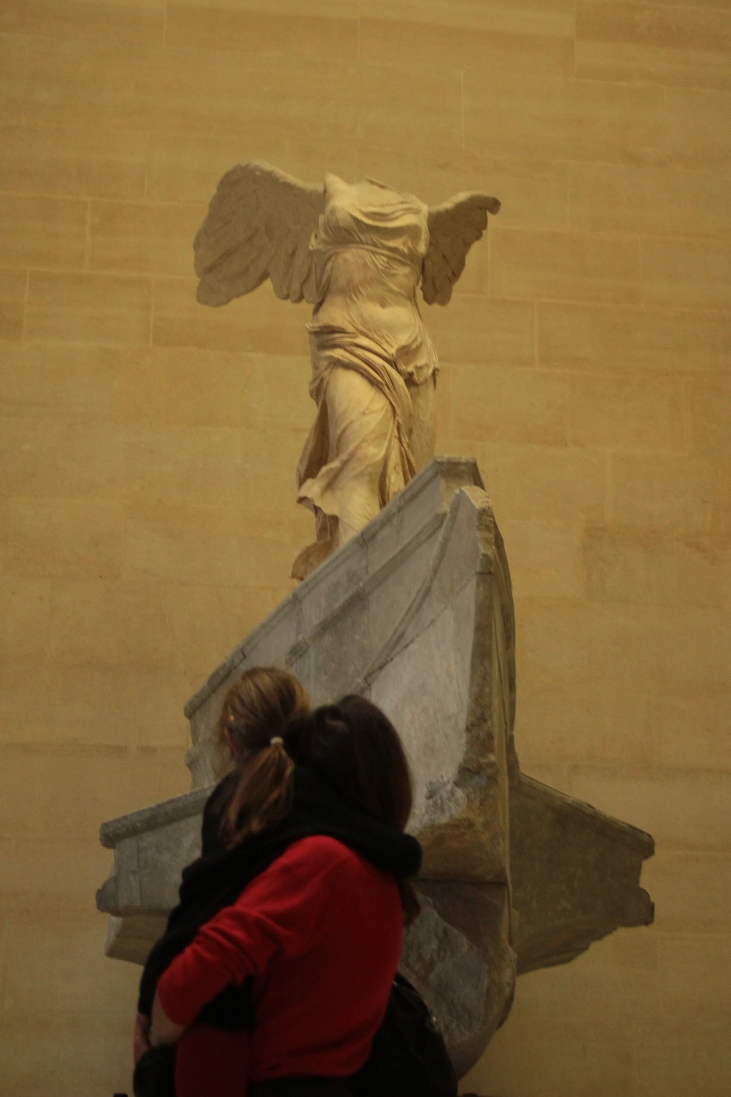 A person with brown hair tied in a ponytail and a red jacket looks up at the ancient marble sculpture of a headless winged figure, displayed against a plain yellow wall.