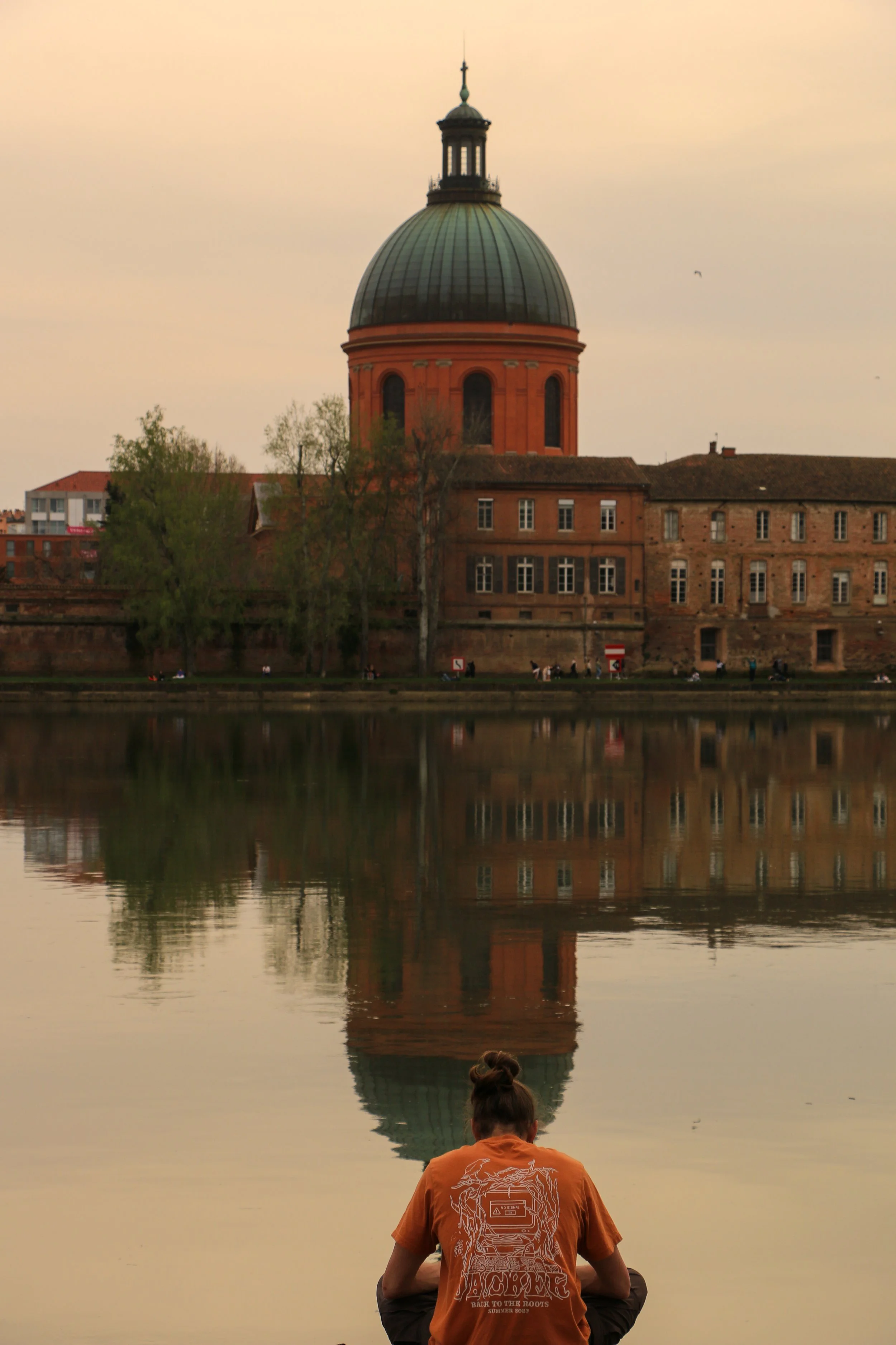 A person sitting by a river with their back to the camera, wearing an orange T-shirt, overlooking a large historic building with a dome, reflected in the water during sunset.