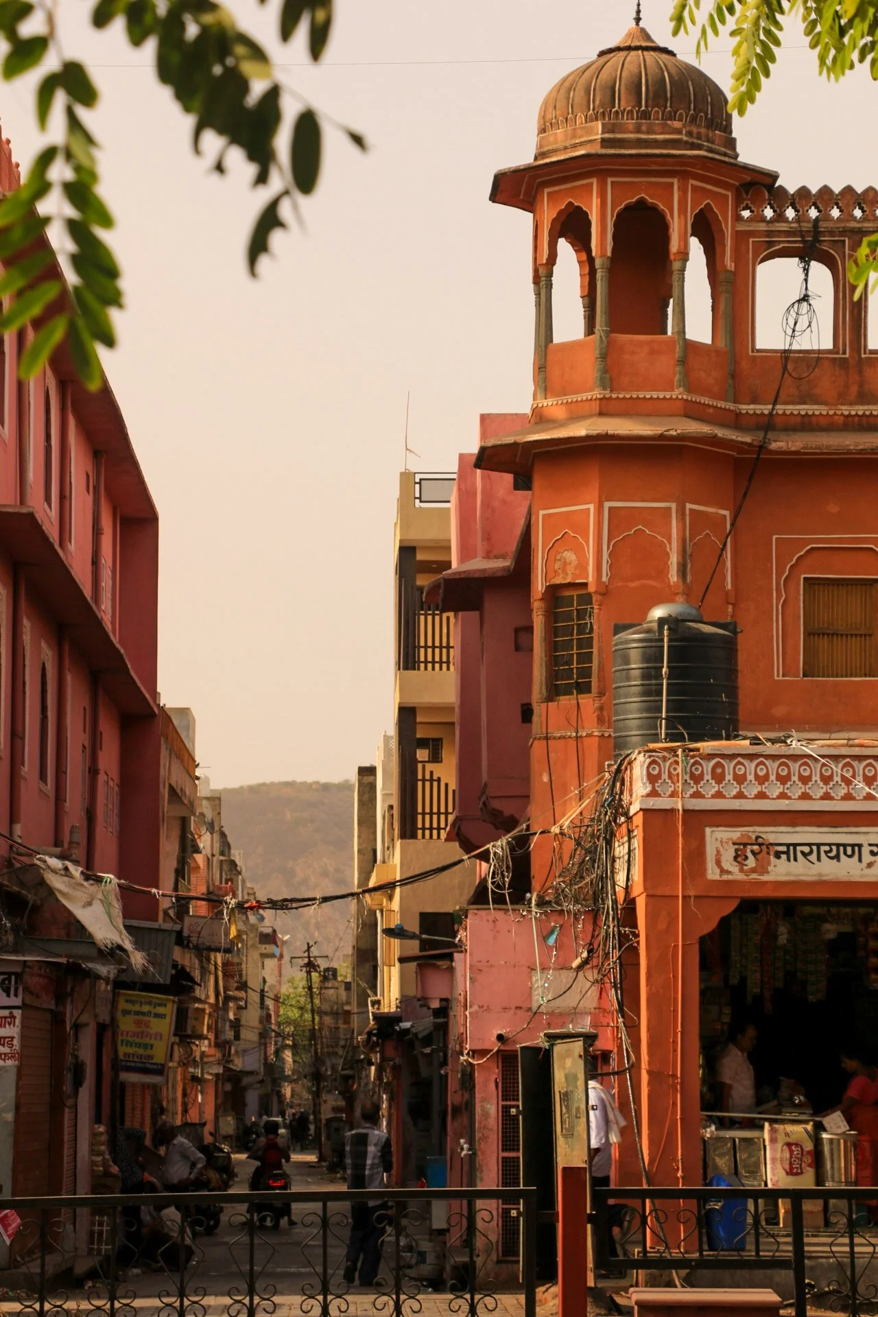 A street scene in India featuring a pink building with traditional architecture on the right, electrical wires hanging across, and people walking or riding scooters in the background with hills in the distance.