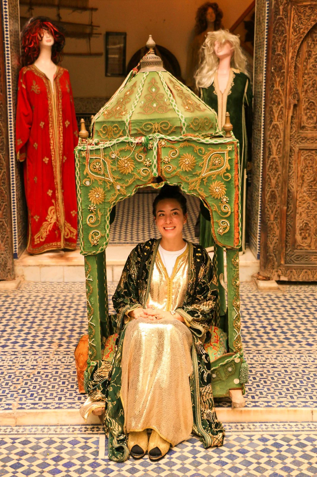 A woman in traditional gold and black clothing sitting under a decorated green canopy, surrounded by mannequins dressed in ornate clothing in a Moroccan-style setting with intricate woodwork and mosaic floor tiles.