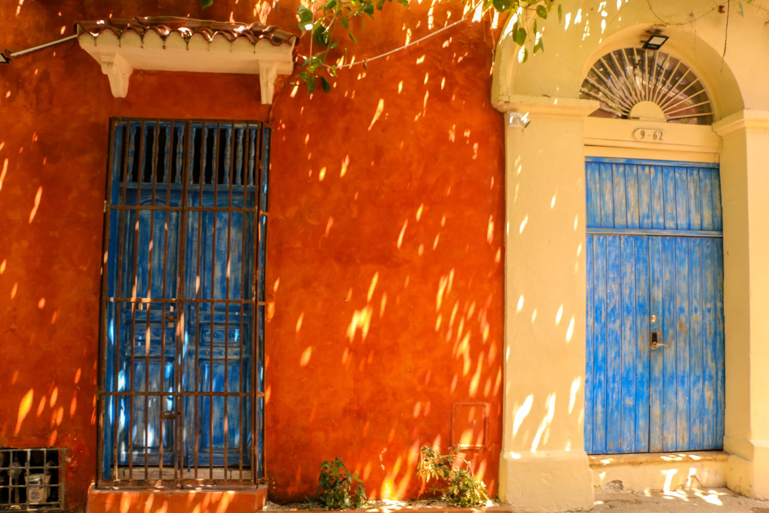 Colorful building façade with orange and yellow walls, blue wooden doors, and decorative elements, with sunlight filtering through leaves casting dappled shadows.