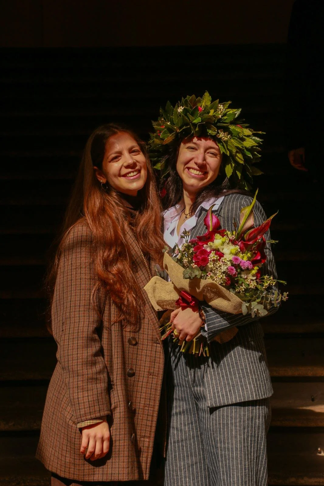 Two women smiling, one wearing a flower crown and holding a bouquet of flowers, with a dark background.