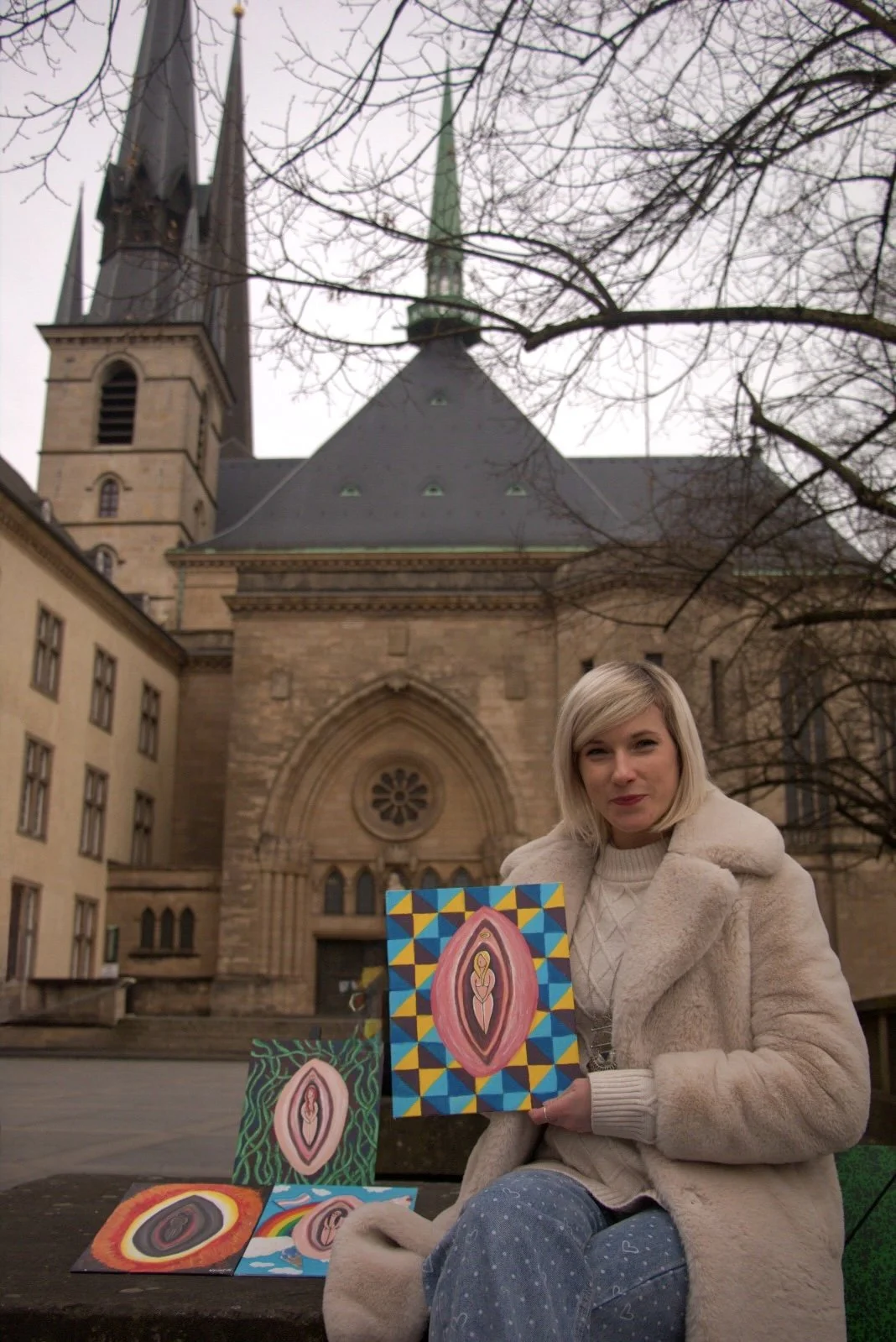 A woman sitting on a bench outdoors, holding a painting of a stylized figure with a colorful background, with other paintings displayed around her, in front of an old church with gothic architecture and spires.