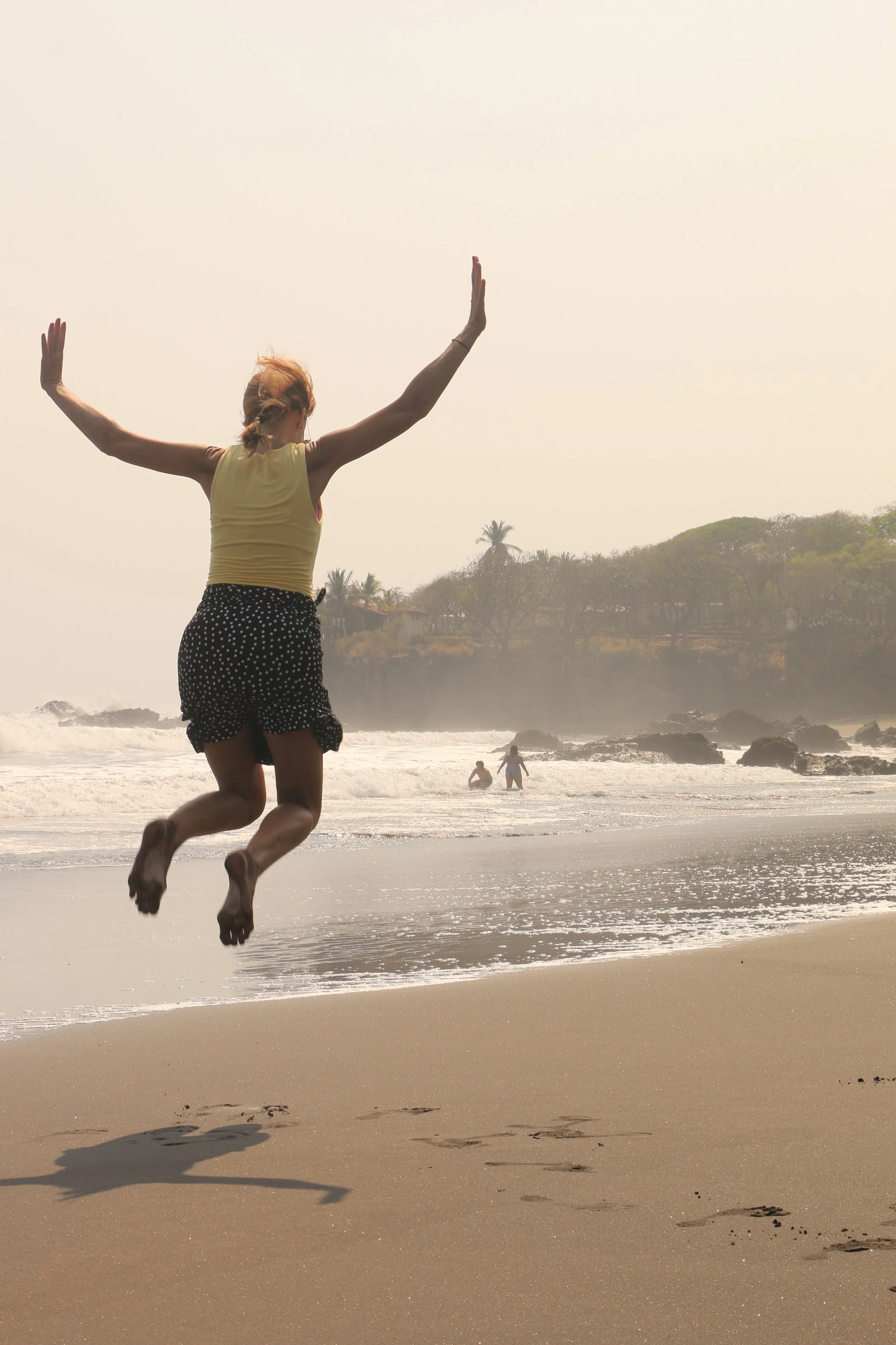 A woman jumping on a beach with her arms outstretched, with ocean waves and palm trees in the background.