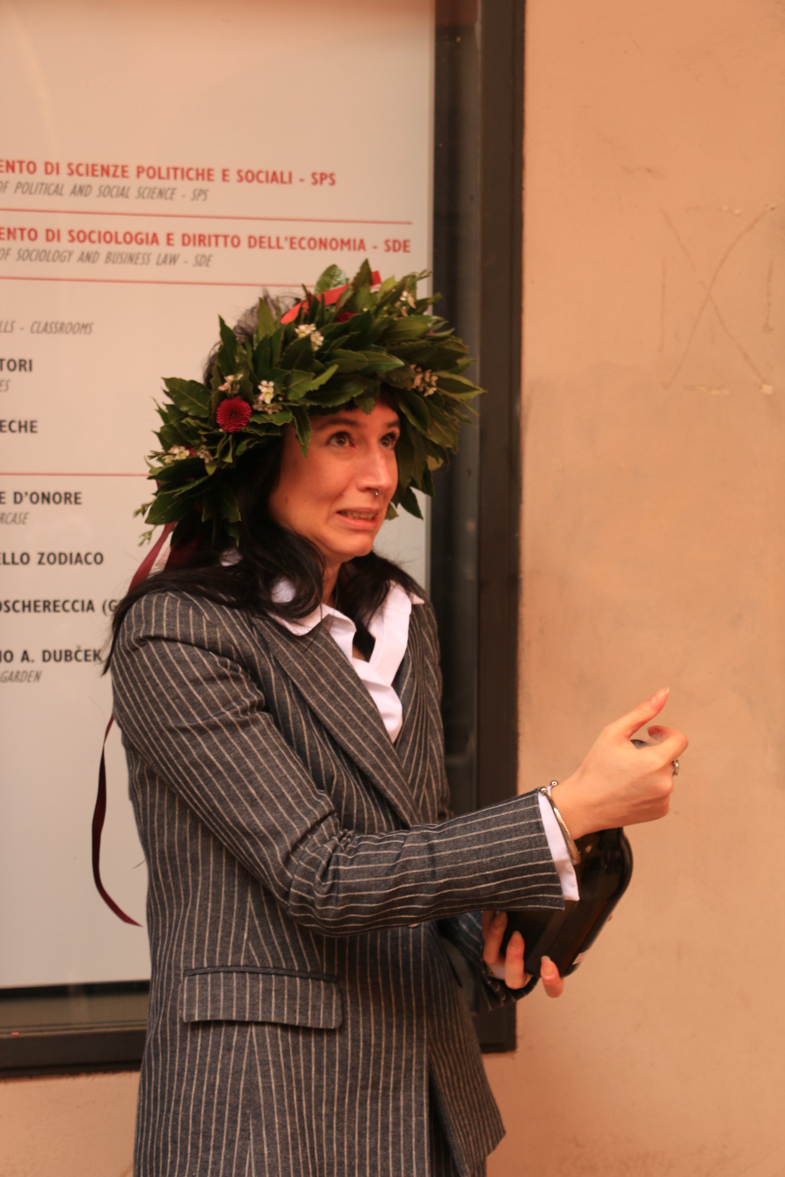 A woman wearing a suit with a floral wreath on her head, making a gesture with her hand.
