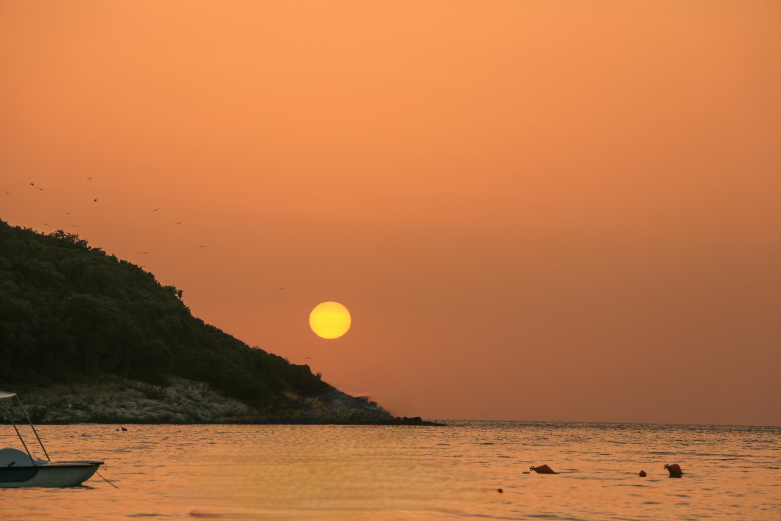 Sunset over a calm sea with a hill on the left, some birds flying, and a small boat floating in the water.