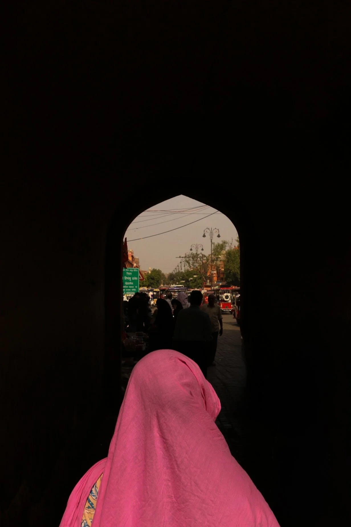 The image shows a person wearing a pink wrap or cloth, sitting or standing in a dark archway, looking out onto a busy street with people, vehicles, and street signs visible in the background outside the arch.
