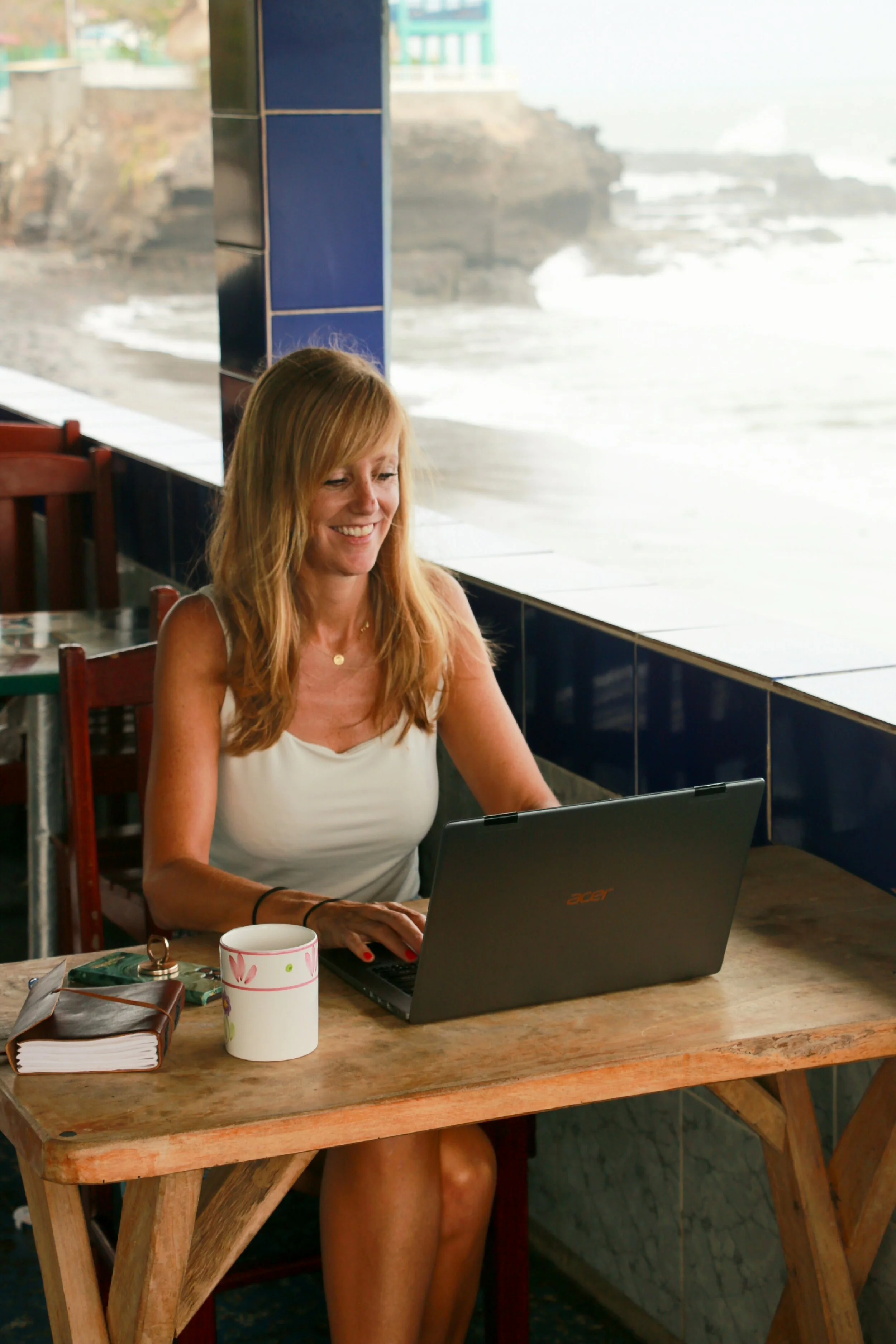 Woman with red hair working on a laptop at a wooden table near large windows overlooking a beach or ocean.