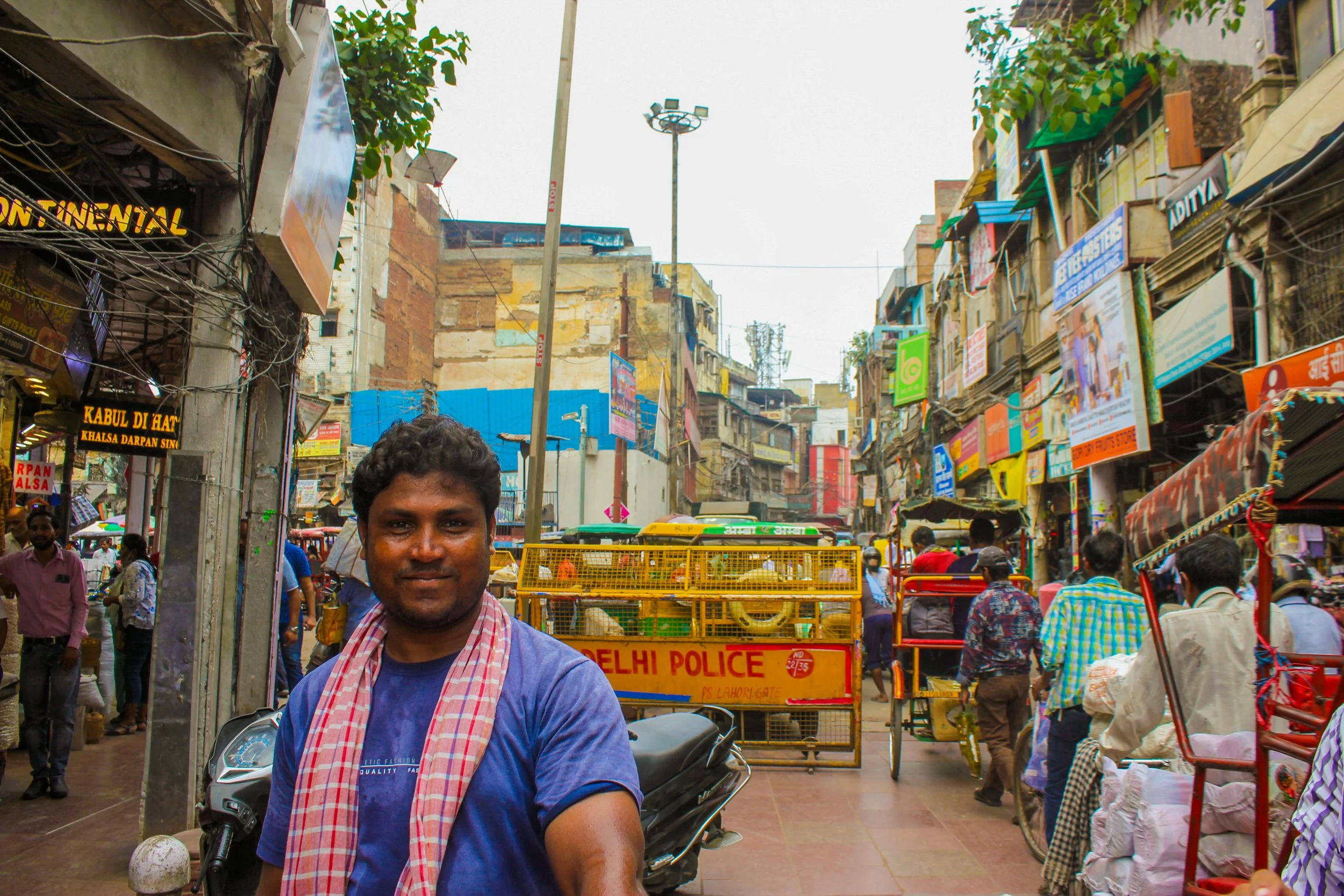 A man smiling in a busy market street with colorful shops and signs, and people walking or riding rickshaws.