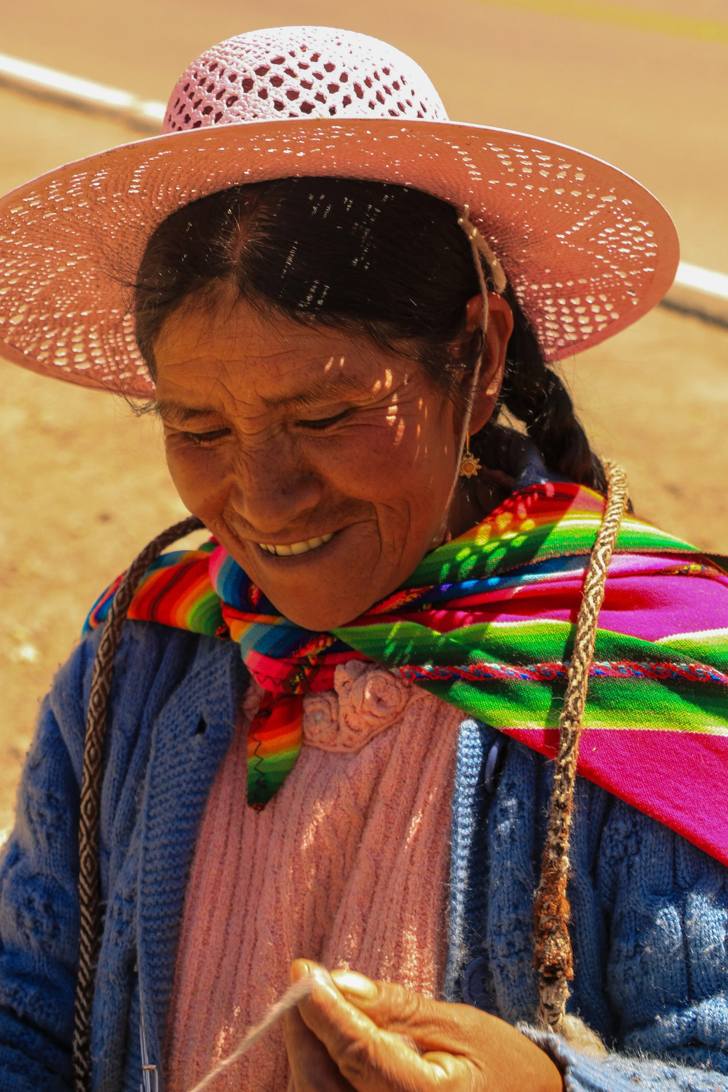 A smiling woman wearing a colorful scarf and a pink straw hat with holes, outside in sunny weather.