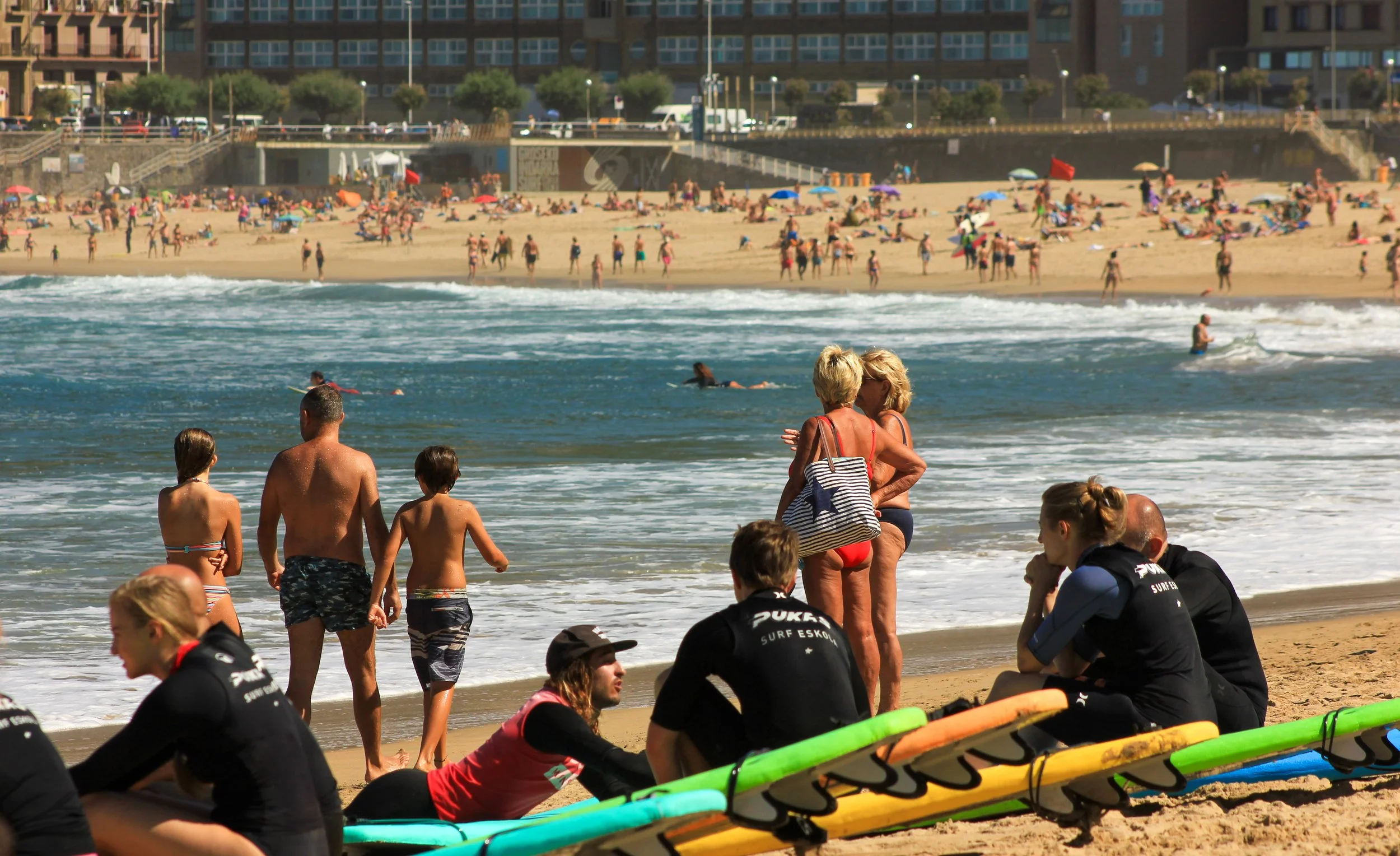 People relaxing and socializing on a busy beach with surfers, children, and others enjoying the sun and sea, with high-rise buildings in the background.