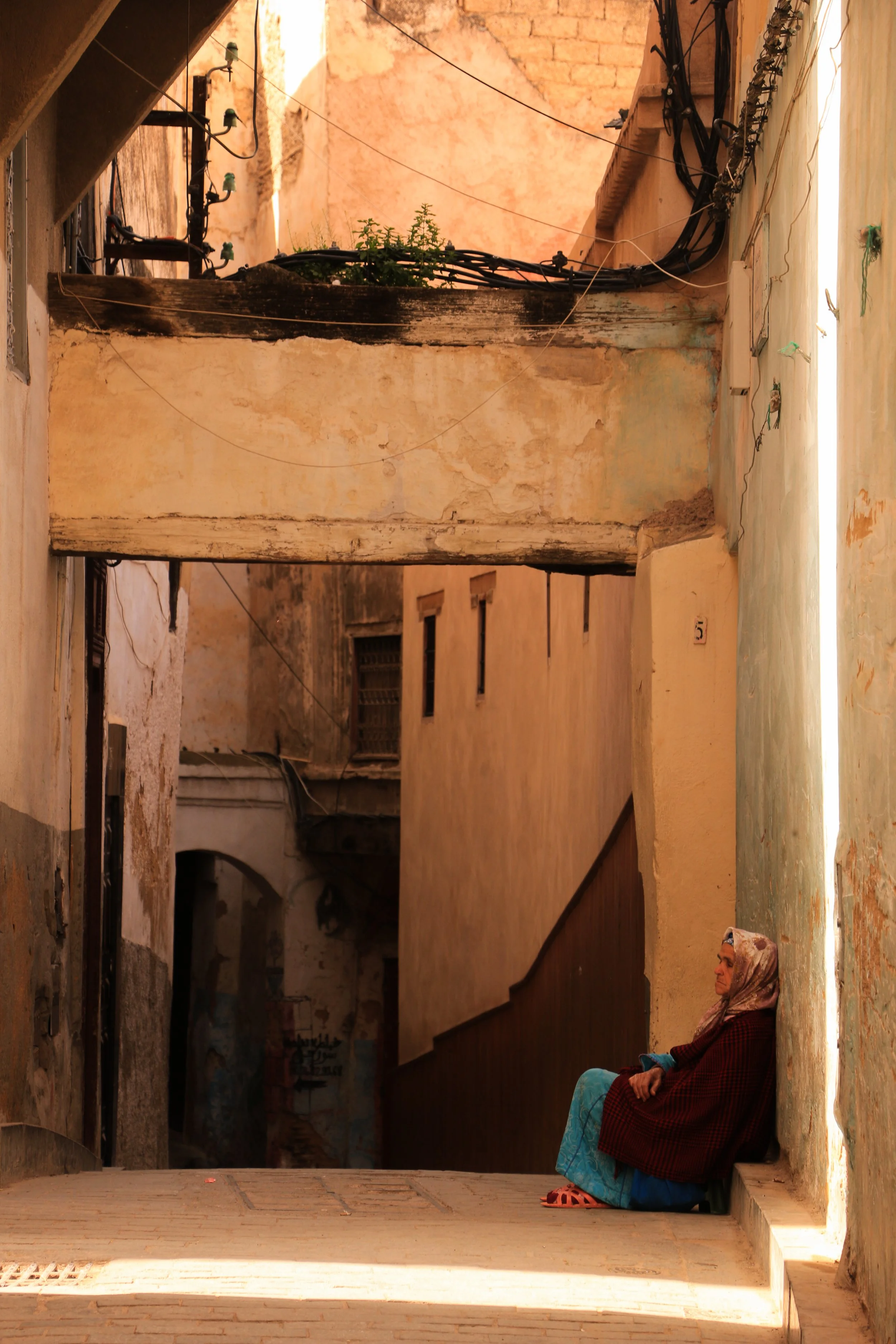 A woman sitting on a step in a narrow, weathered alley with aged walls and overhanging beams, wearing a headscarf and colorful clothing.