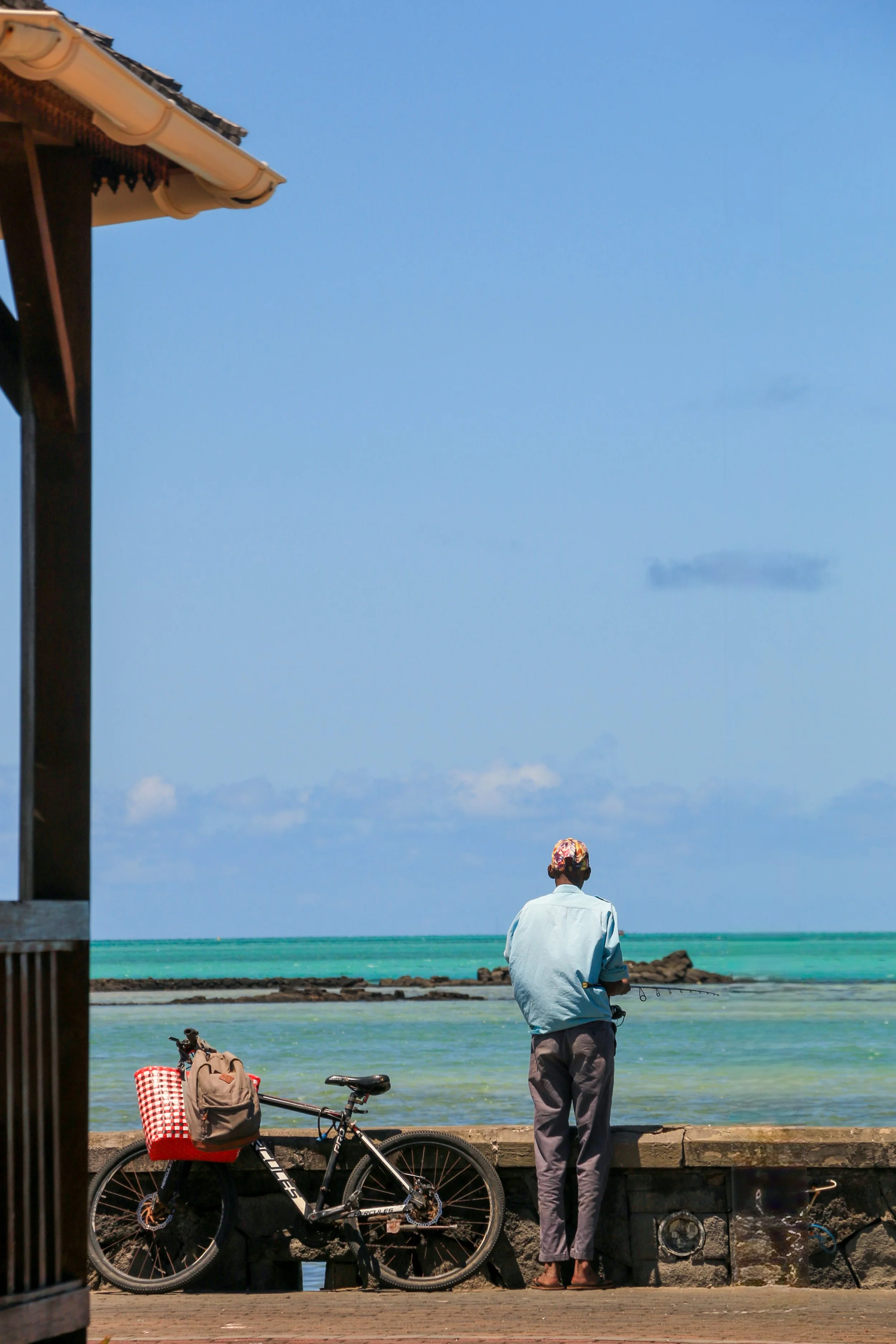 A man standing on a seaside walkway, fishing, with a bicycle loaded with a backpack and checkered bag resting against it, near a building under a clear blue sky.
