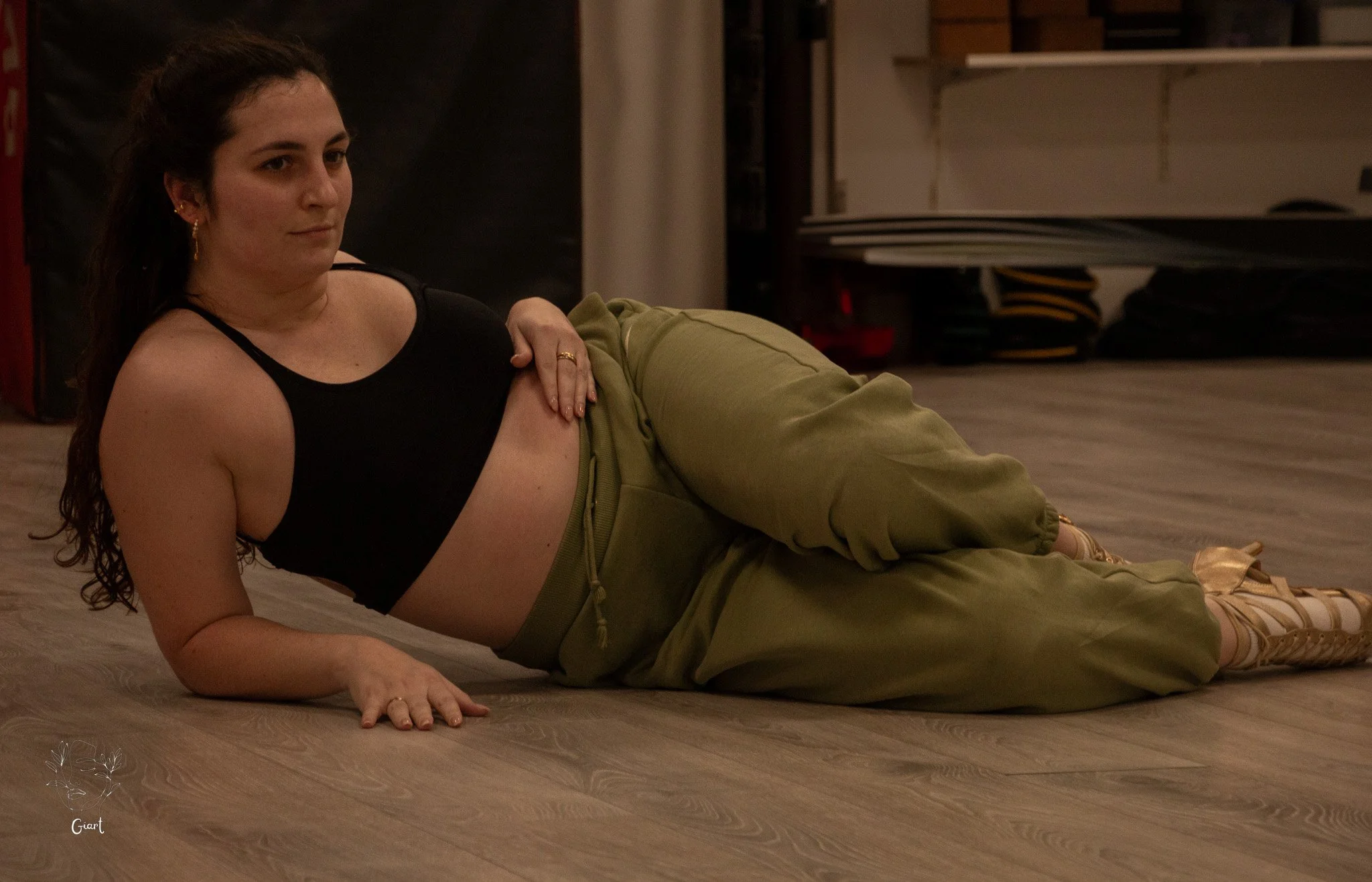 Woman lying on a wooden floor, supporting herself on her side with her left arm, wearing a black crop top and loose green pants.