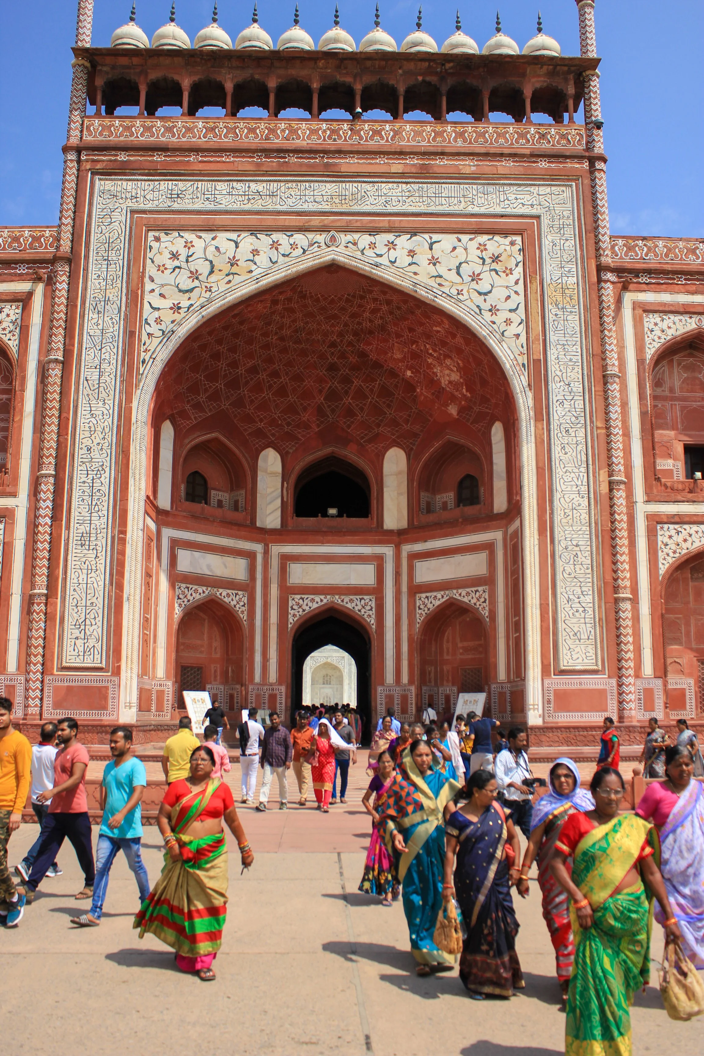 People walking in front of a Mughal-era red sandstone gateway with intricate white marble inlay and Islamic calligraphy, part of a historic monument.