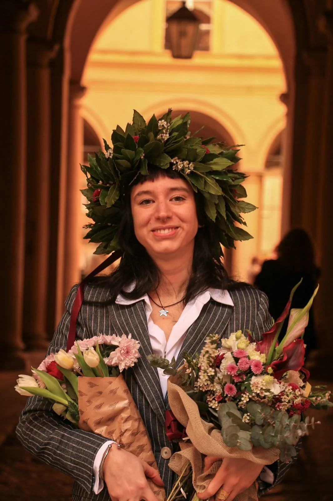 A woman with dark hair and a floral wreath on her head, holding multiple bouquets of flowers, smiling indoors.