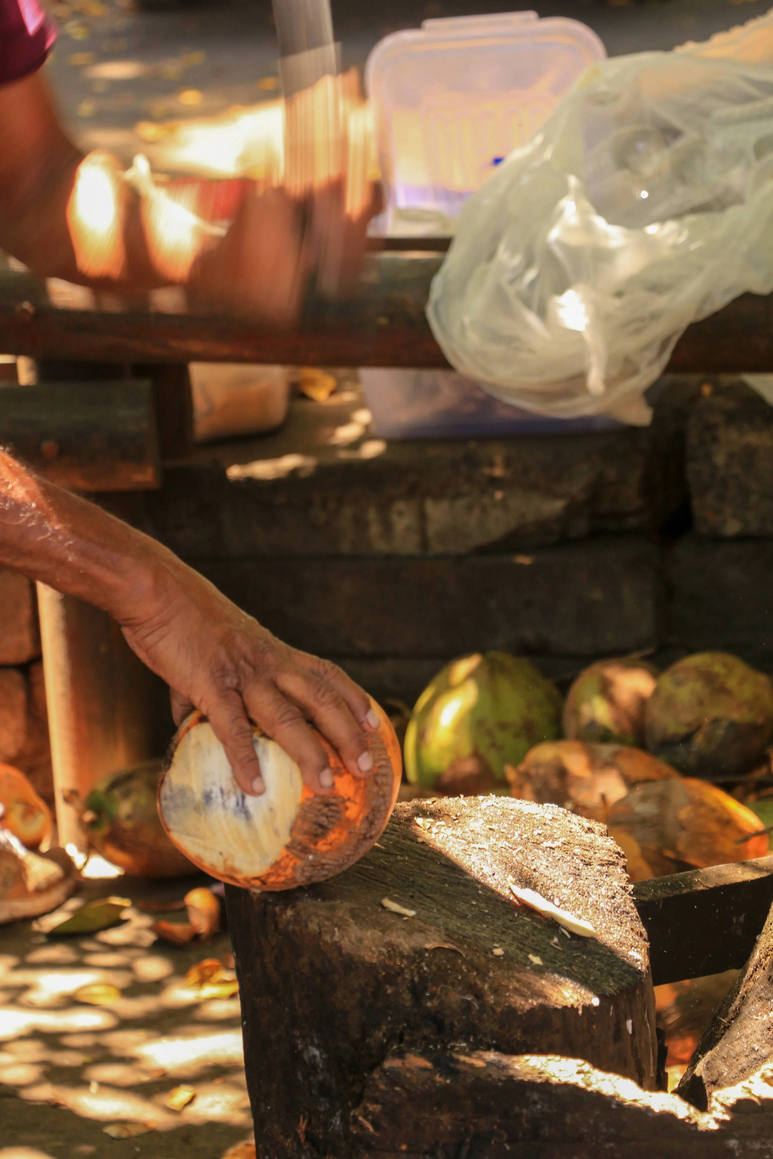 Close-up of a person cracking open a coconut using a large knife on a wooden chopping block. Several avocados are visible in the background and a blurred table with a plastic bag and containers.
