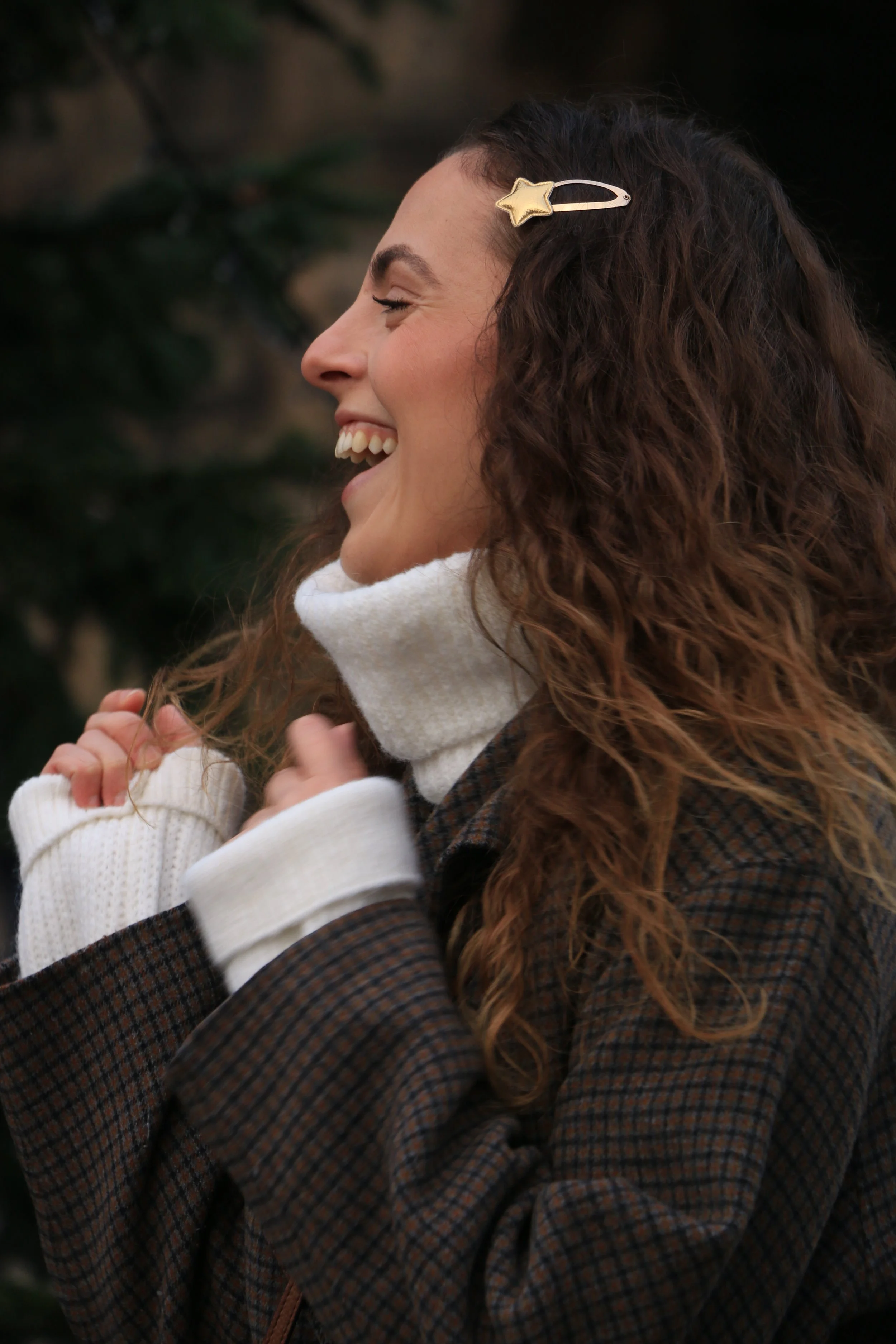 A woman with curly brown hair wearing a star-shaped hair clip, white turtleneck, and checkered jacket, smiling and enjoying a moment outdoors.