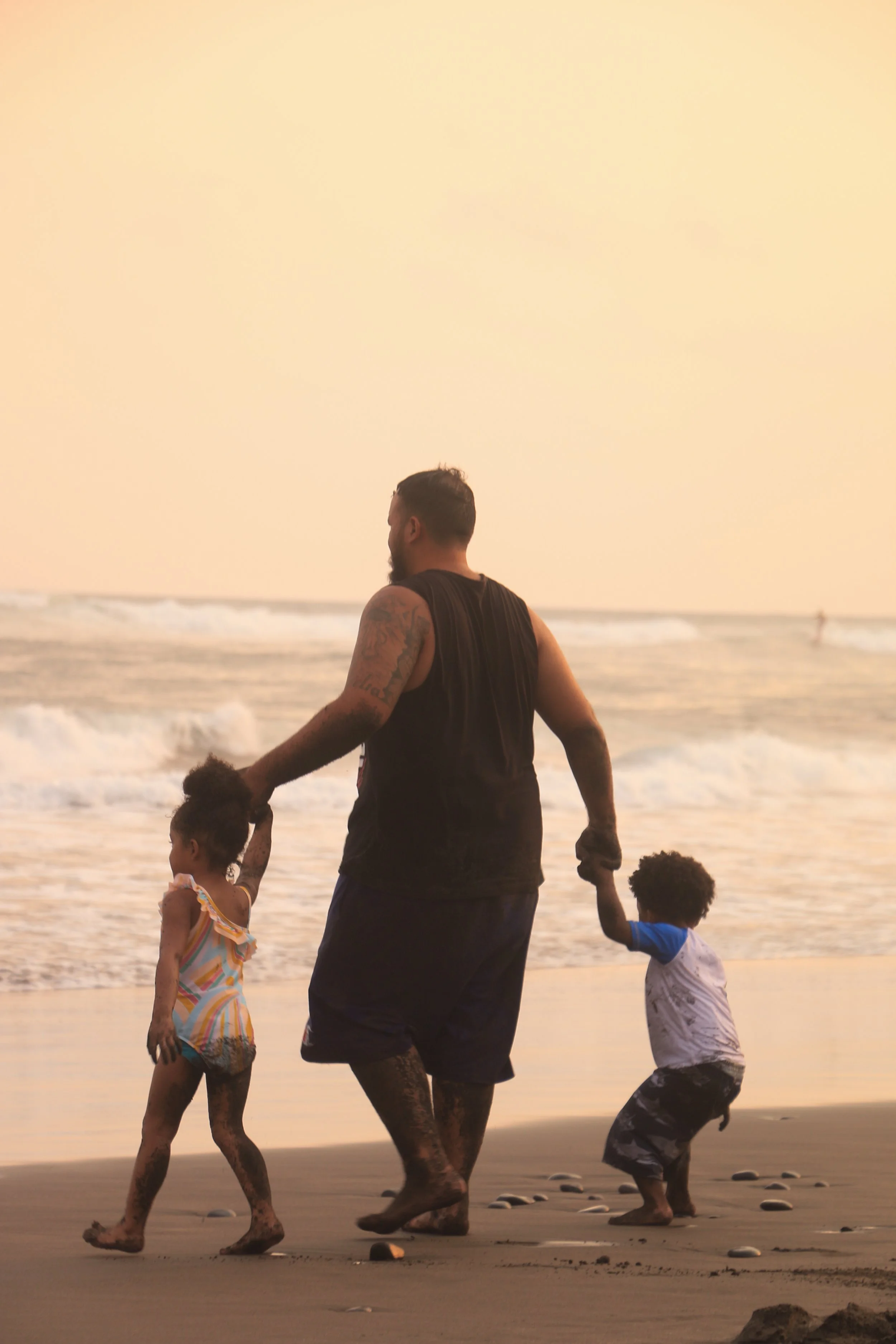 A man holding hands with two children walks along the beach at sunset, with ocean waves in the background.