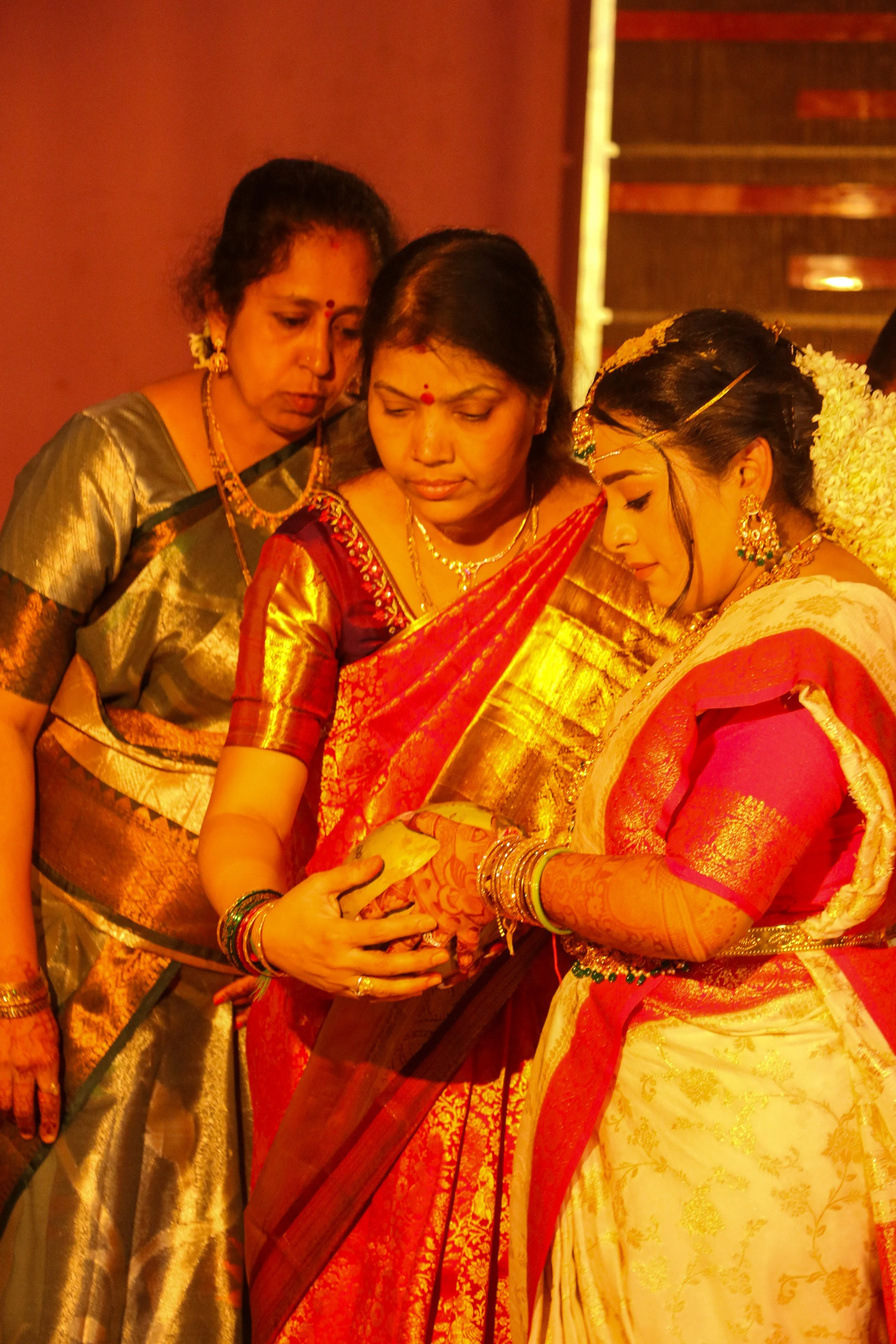 Four women dressed in traditional Indian sarees, gathered closely together, looking at a piece of paper or document, during a cultural or religious event.