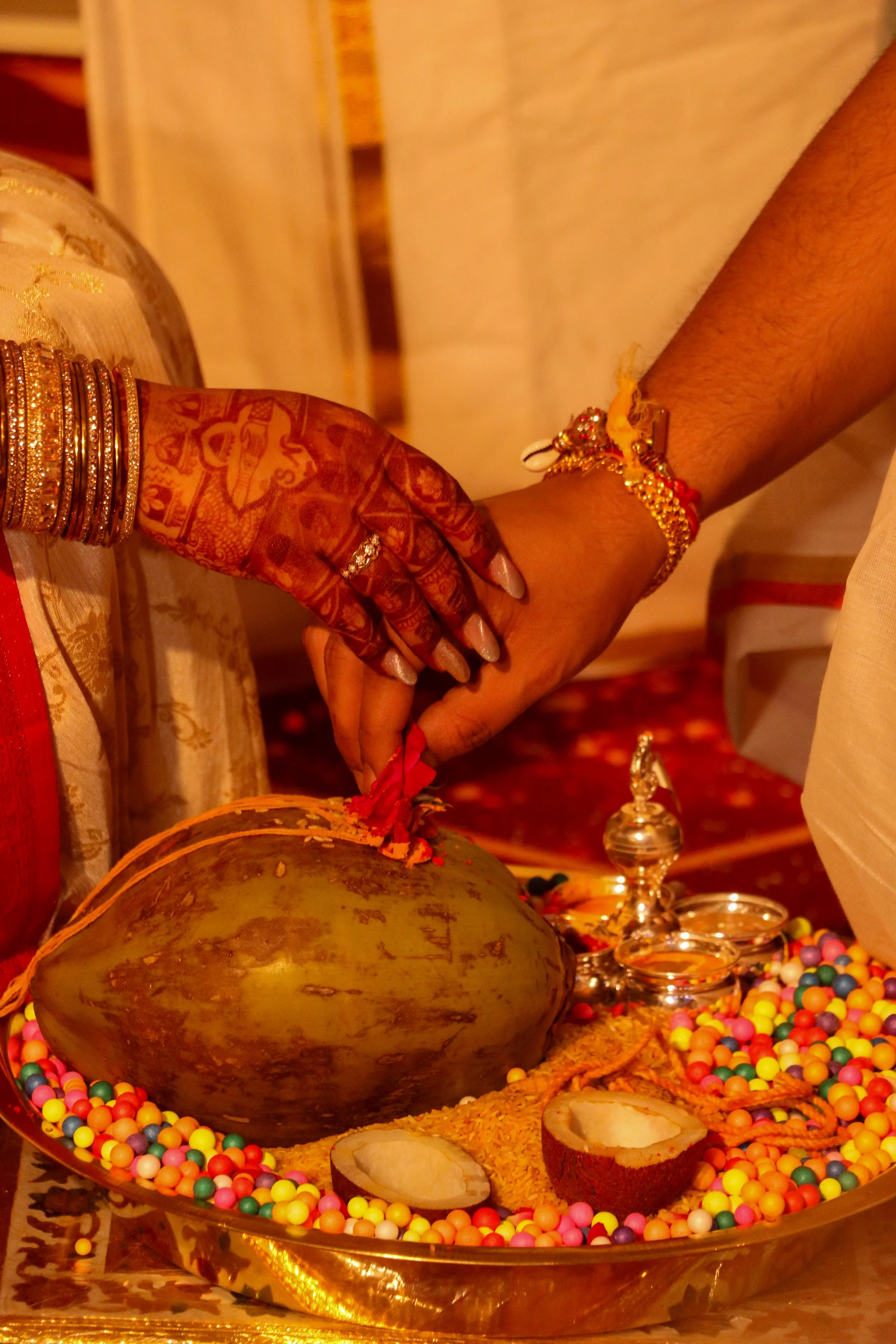 A traditional Indian wedding ritual with one person placing a red mark on a coconut on a decorated tray containing colorful beads, a coconut with its husk partially peeled, small metal containers, and a silver ornament.