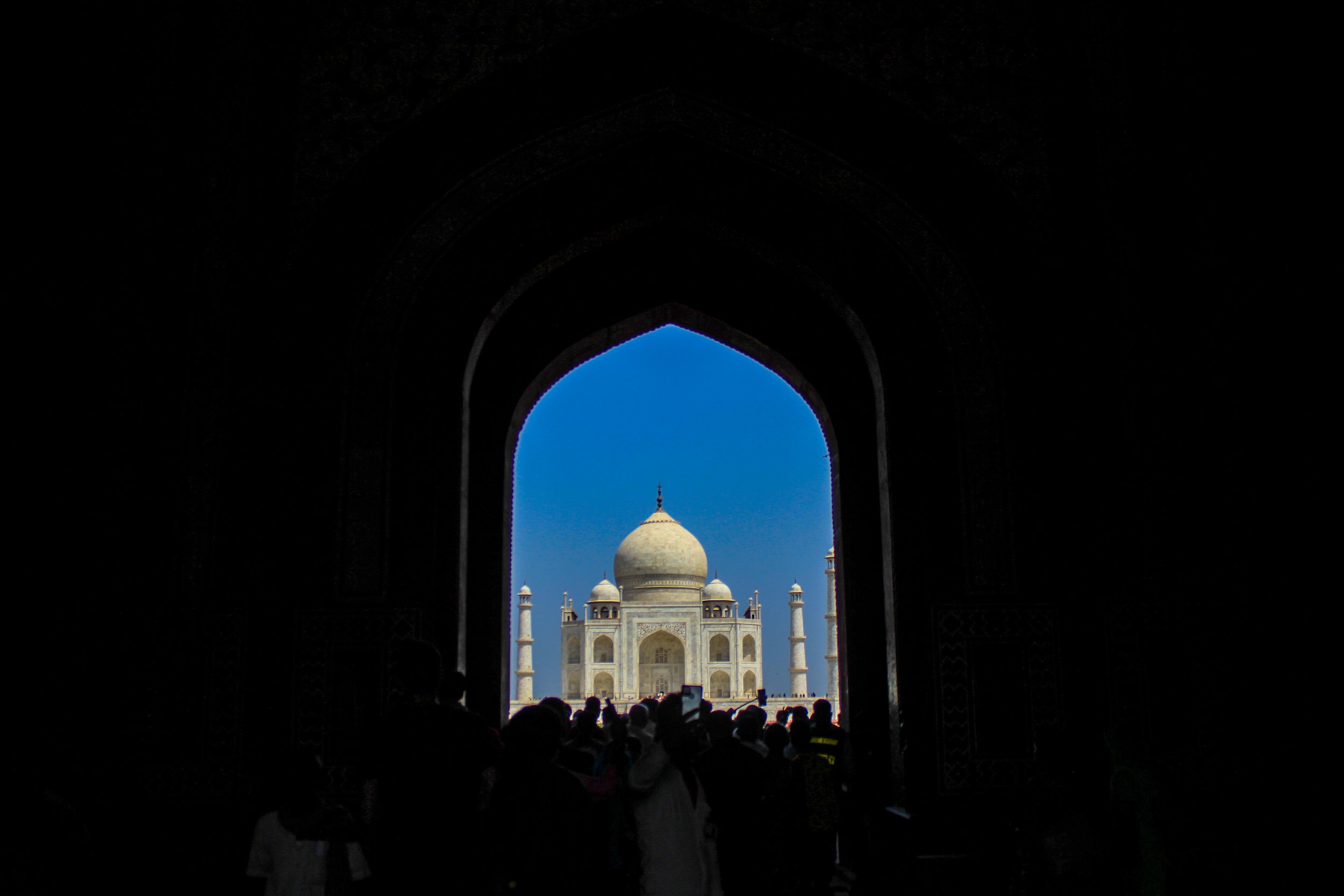 View of the Taj Mahal through a dark arched entrance, with visitors silhouetted against a bright blue sky.