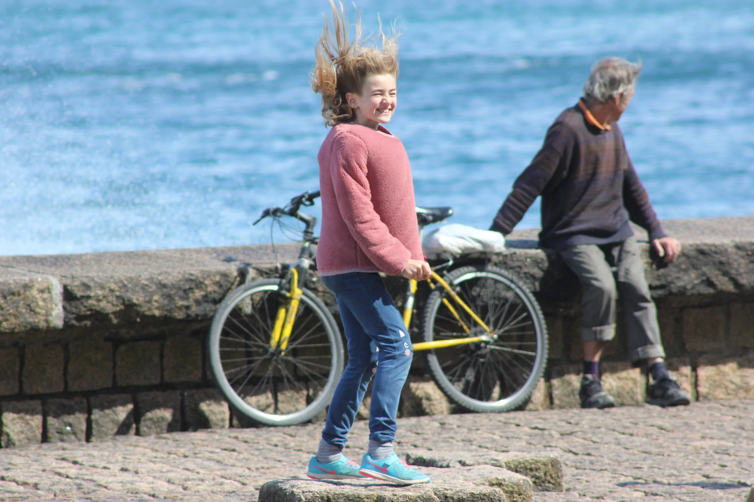 A young girl with long, wavy blonde hair, wearing a pink fuzzy sweater, blue jeans with patches, and teal sneakers, smiling and jumping in front of a coastal scene. Behind her, an older man with gray hair, wearing a dark sweater and gray pants, sits on a stone wall by the water. A yellow bicycle leans against the wall next to him, with the ocean visible in the background.