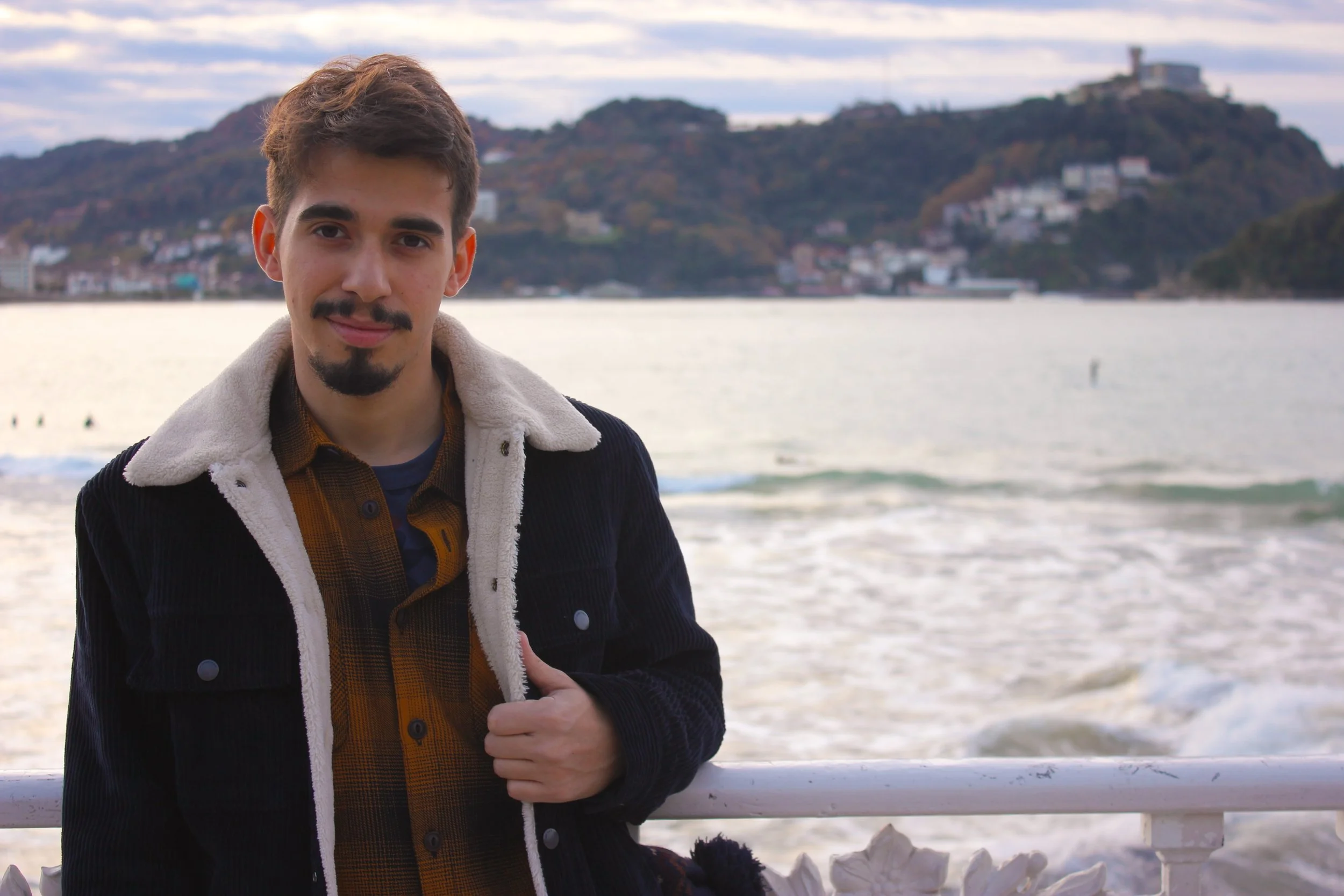 A young man with brown hair and a beard standing by a railing at the beach during sunset, with ocean waves and hills with buildings in the background.