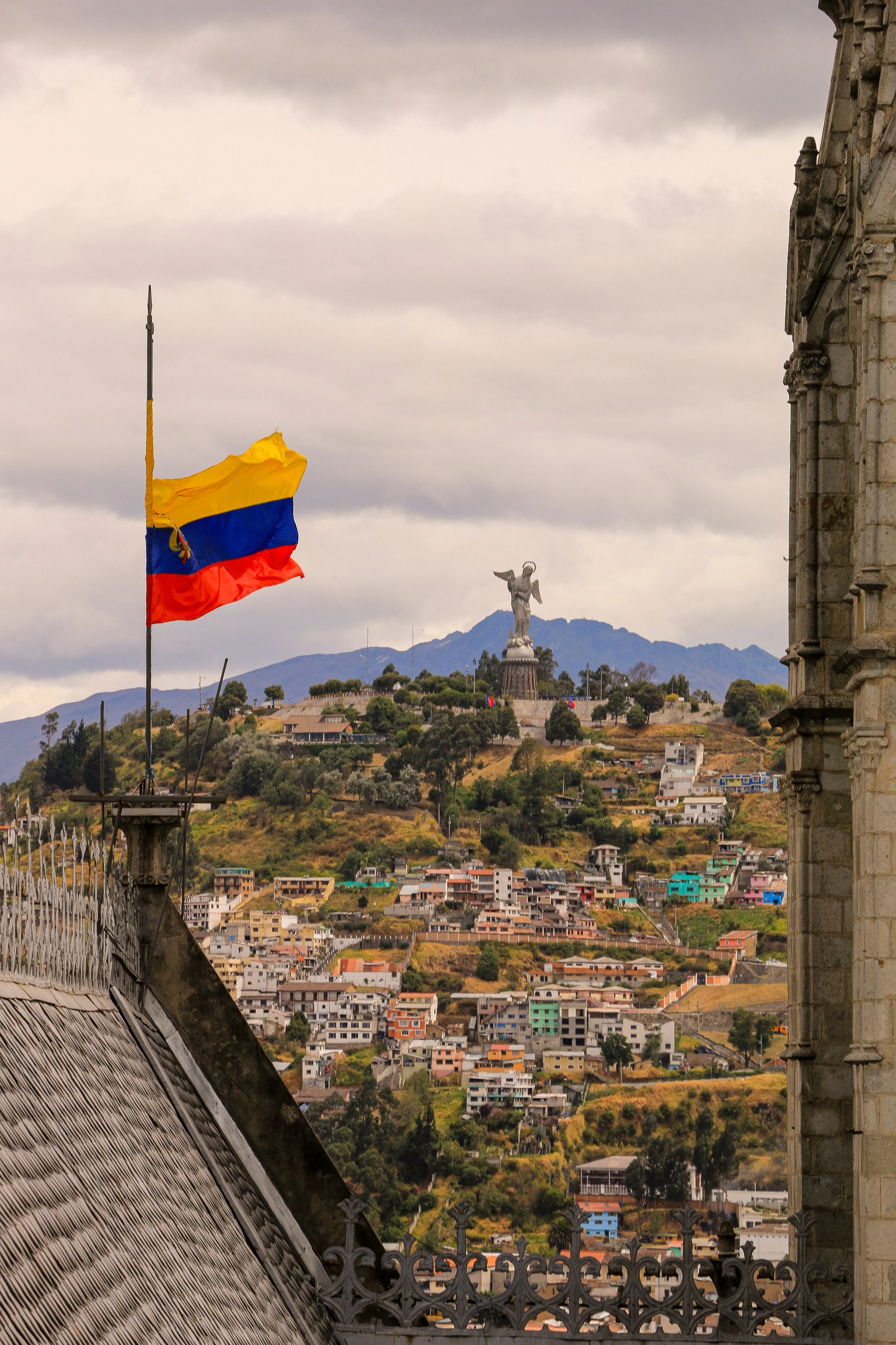 View of a hillside with colorful houses, a large Statue of Liberty-like statue on top, and a mounted Ecuadorian flag in the foreground.