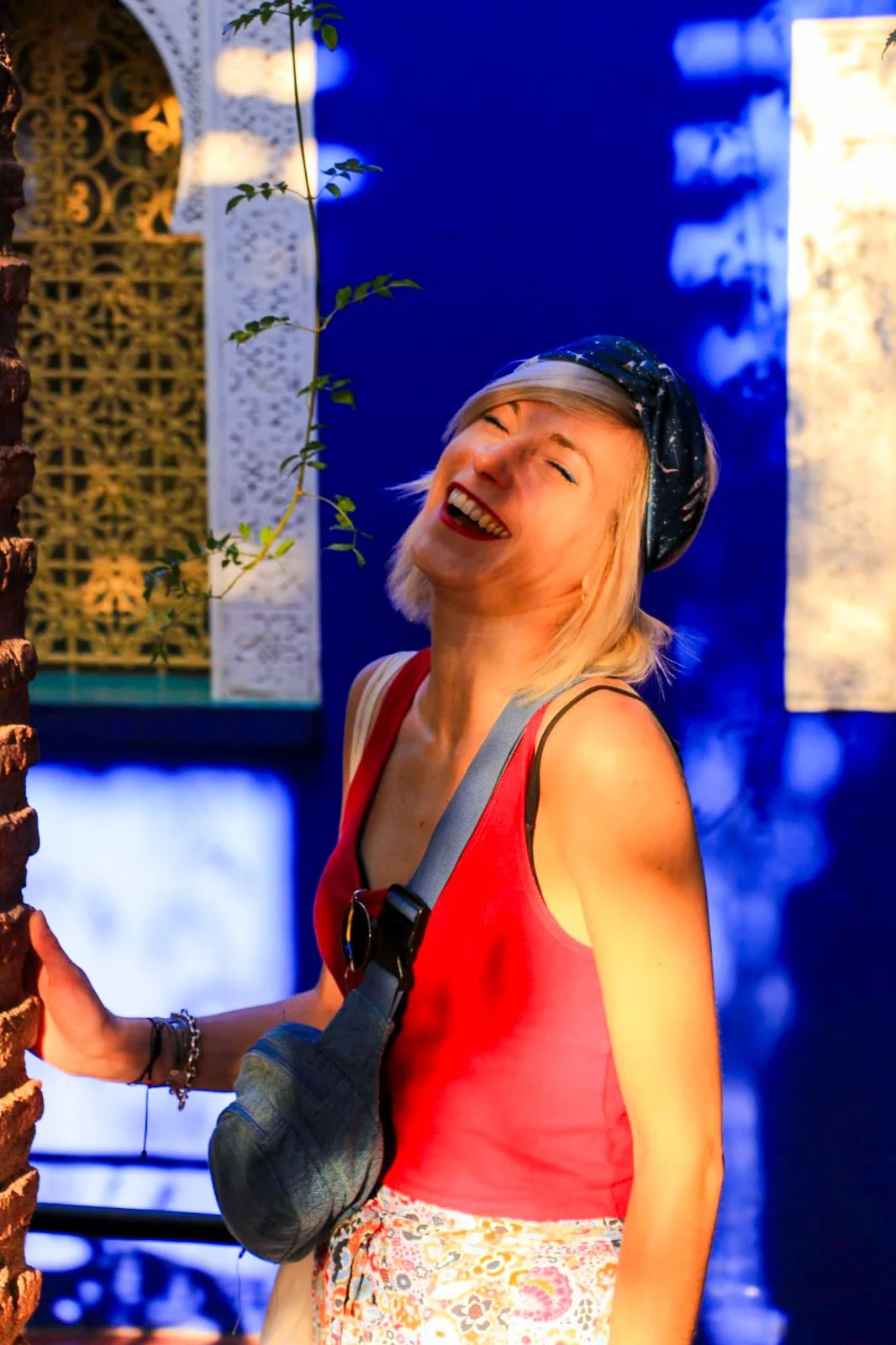 A woman with blonde hair wearing a blue headscarf, a red sleeveless top, and patterned pants, smiling joyfully with her eyes closed. She's standing outdoors near a brick column, with lush green vines and a decorative lattice in the background, lit by warm yellow and cool blue lighting.