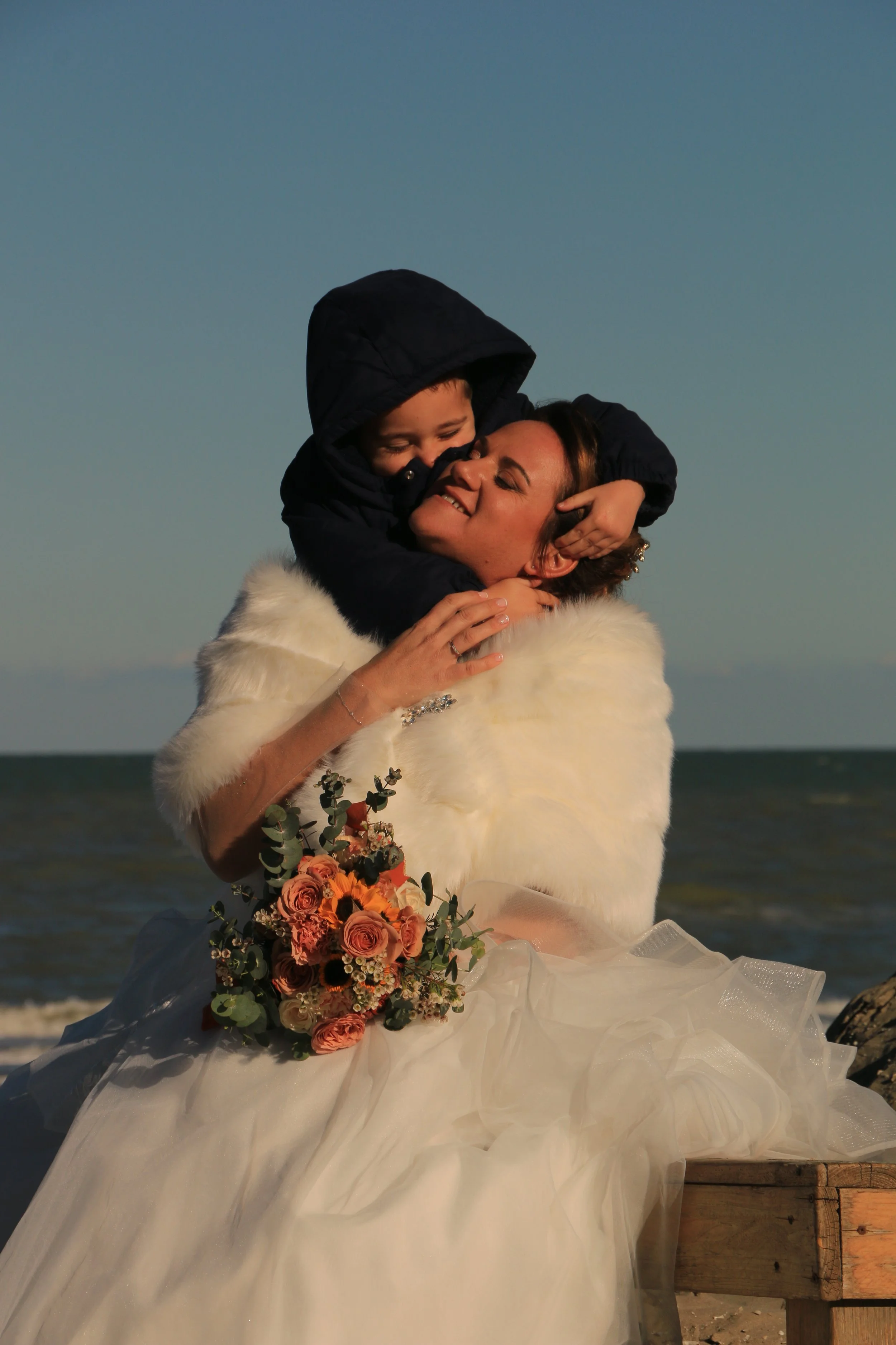 A woman in a wedding dress and furry white shawl hugging a boy in a dark hoodie at the beach, holding a bouquet of pink and orange flowers, with ocean in the background.